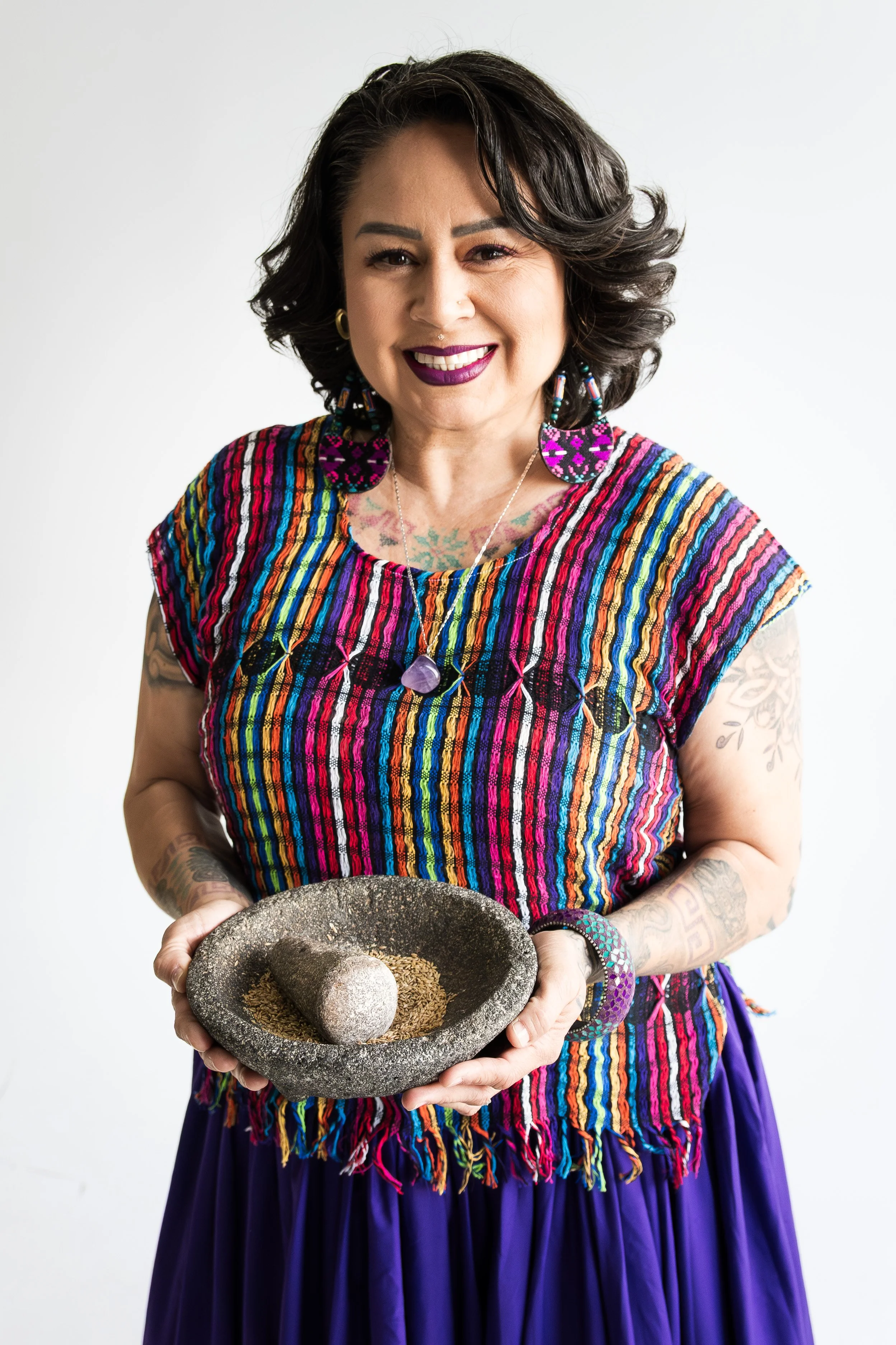 Woman with dark wavy hair, purple lipstick, colorful striped top, holding a stone mortar with herbs and two pestle stones, against a plain white background.