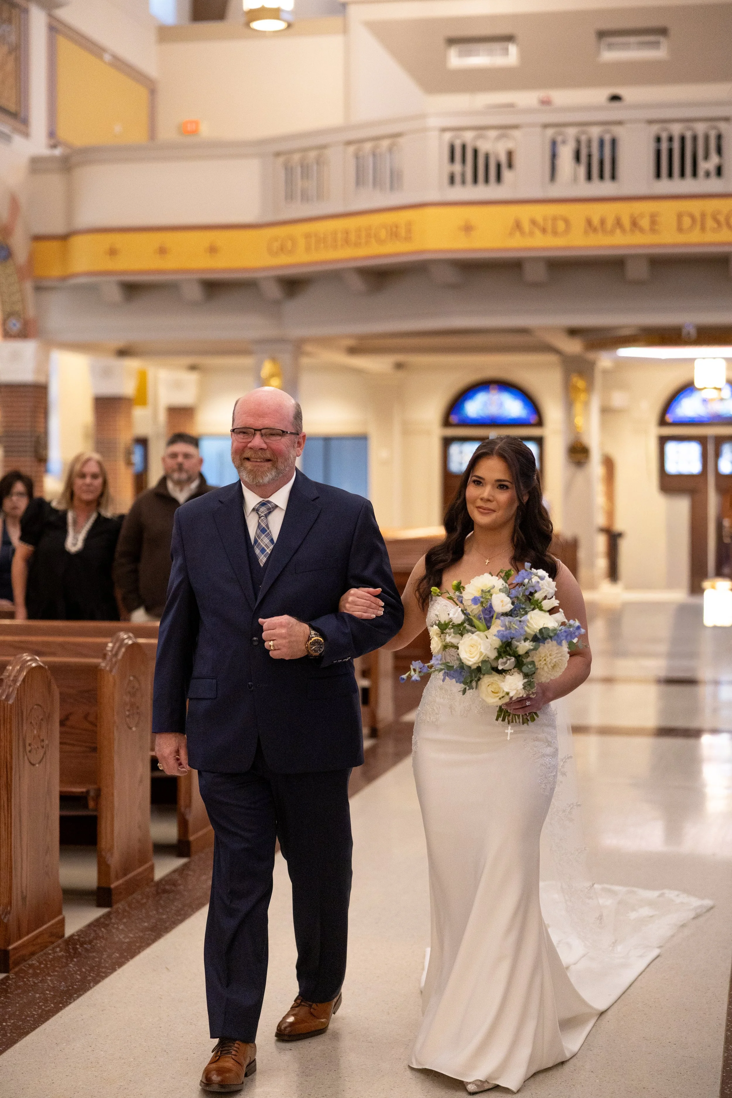 Bride walking down the aisle with her father during a church wedding ceremony, wearing a white gown and holding a bouquet of white and purple flowers captured by Mandy Laine Photo a College Station wedding photographer