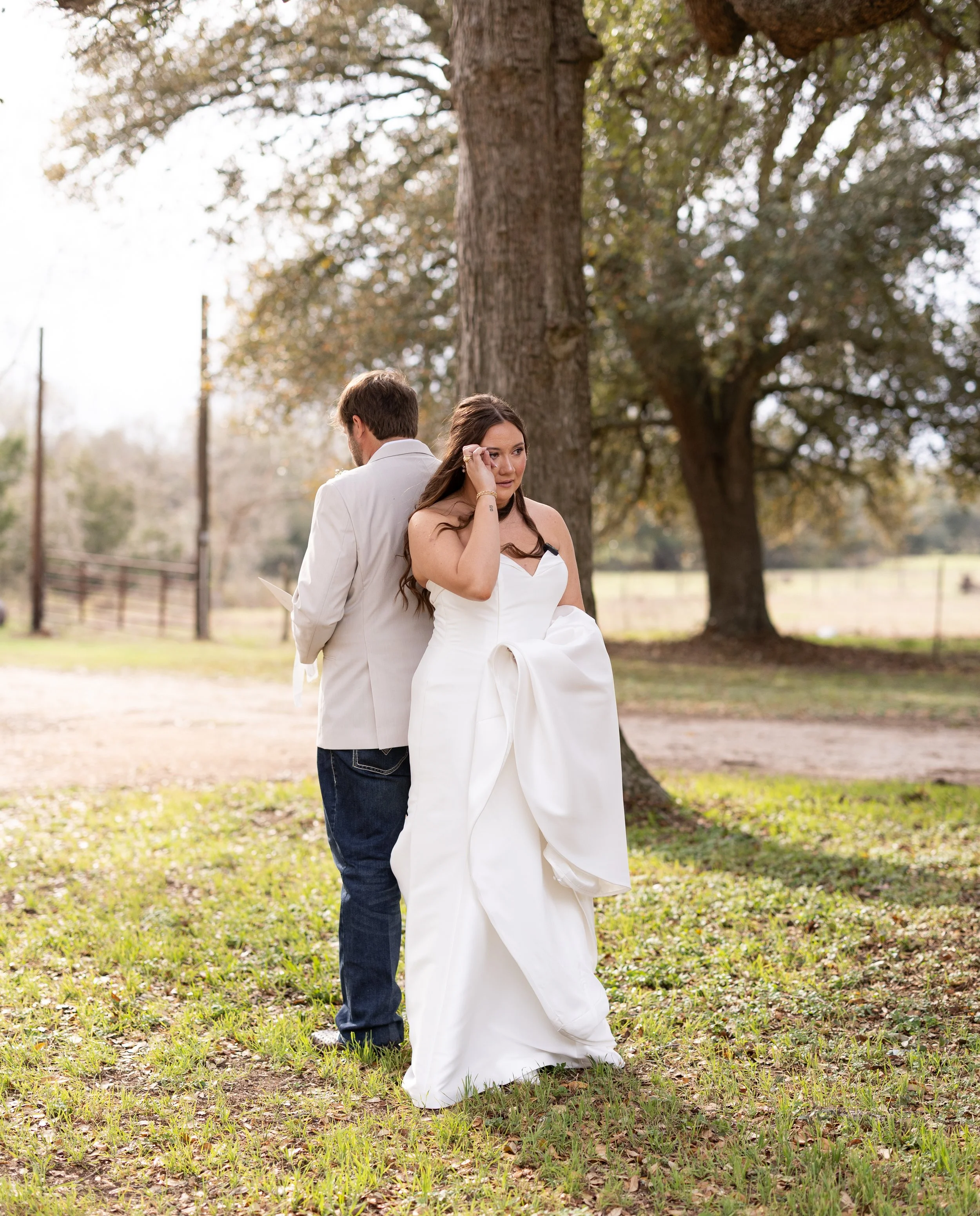 Bride in a white wedding dress holding her gown outdoors next to a large tree, with groom in a light-colored suit reading a paper in a rural setting with trees and a fence captured by College Station wedding photographer Mandy Laine Photo