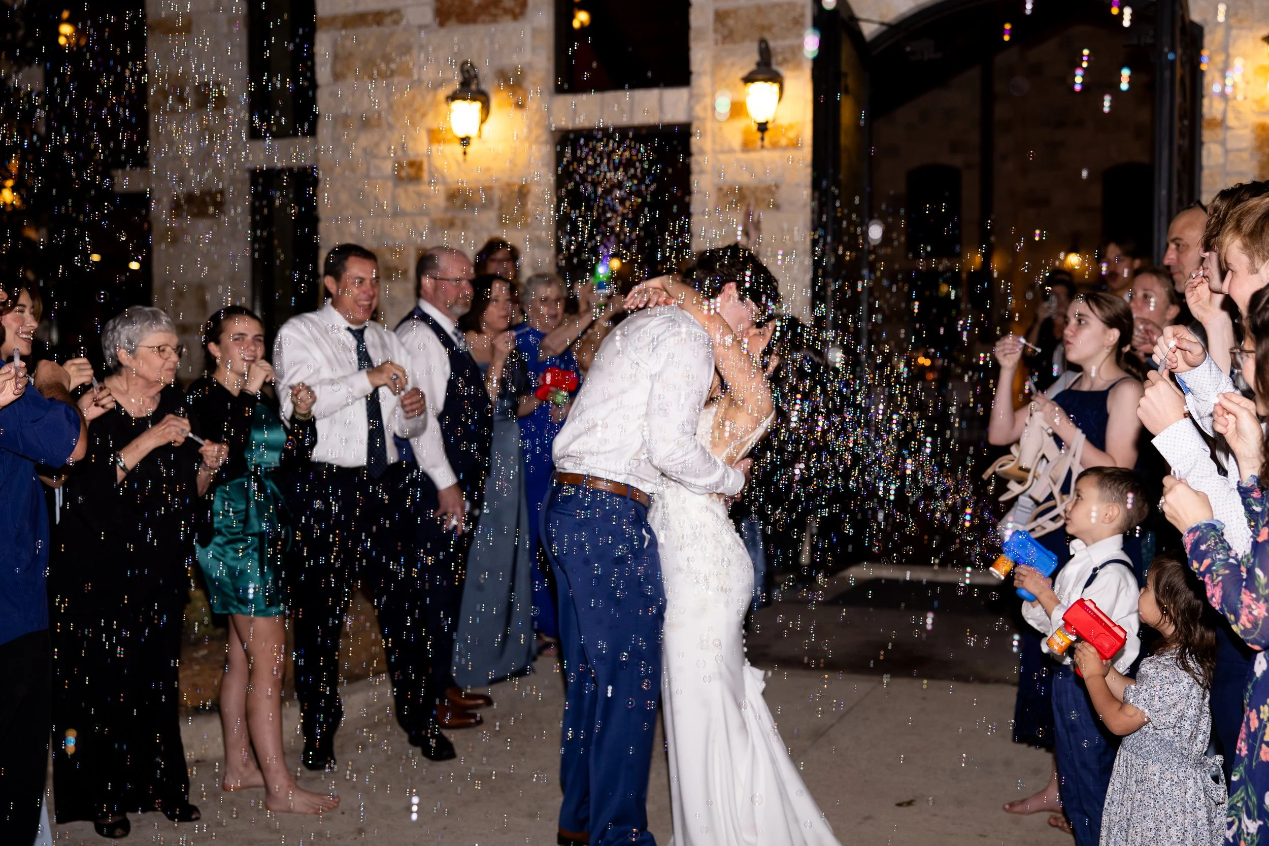 Bride and groom sharing a kiss under bubbles at night, surrounded by wedding guests outside a stone building with warm lights. Captured in natural light to highlight a joyful, romantic wedding celebration. Captured by Mandy Laine Photo, a college sta
