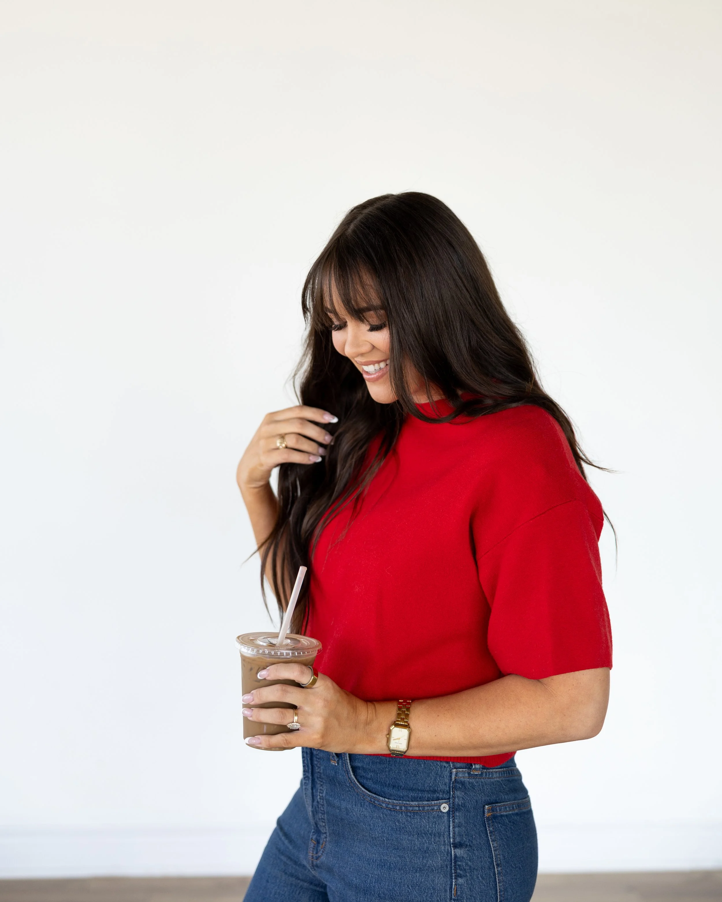 Smiling woman with long dark hair in a red shirt and blue jeans holding an iced coffee indoors.