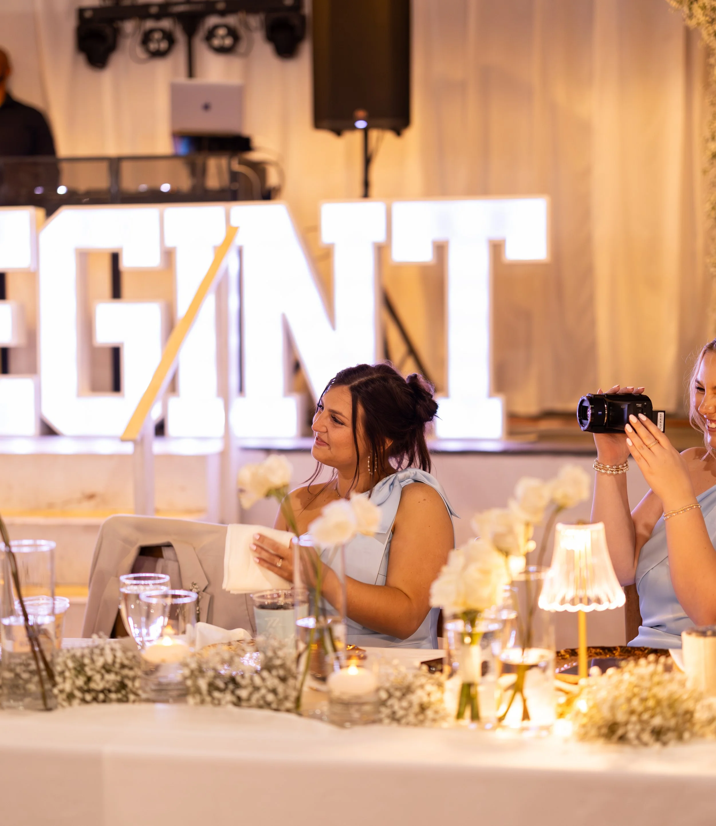 A woman at a wedding reception table, smiling and holding a napkin, with a woman on her right taking a photo with a camera, and large illuminated letters in the background. Captured by Mandy Laine Photo, College Station wedding photographer