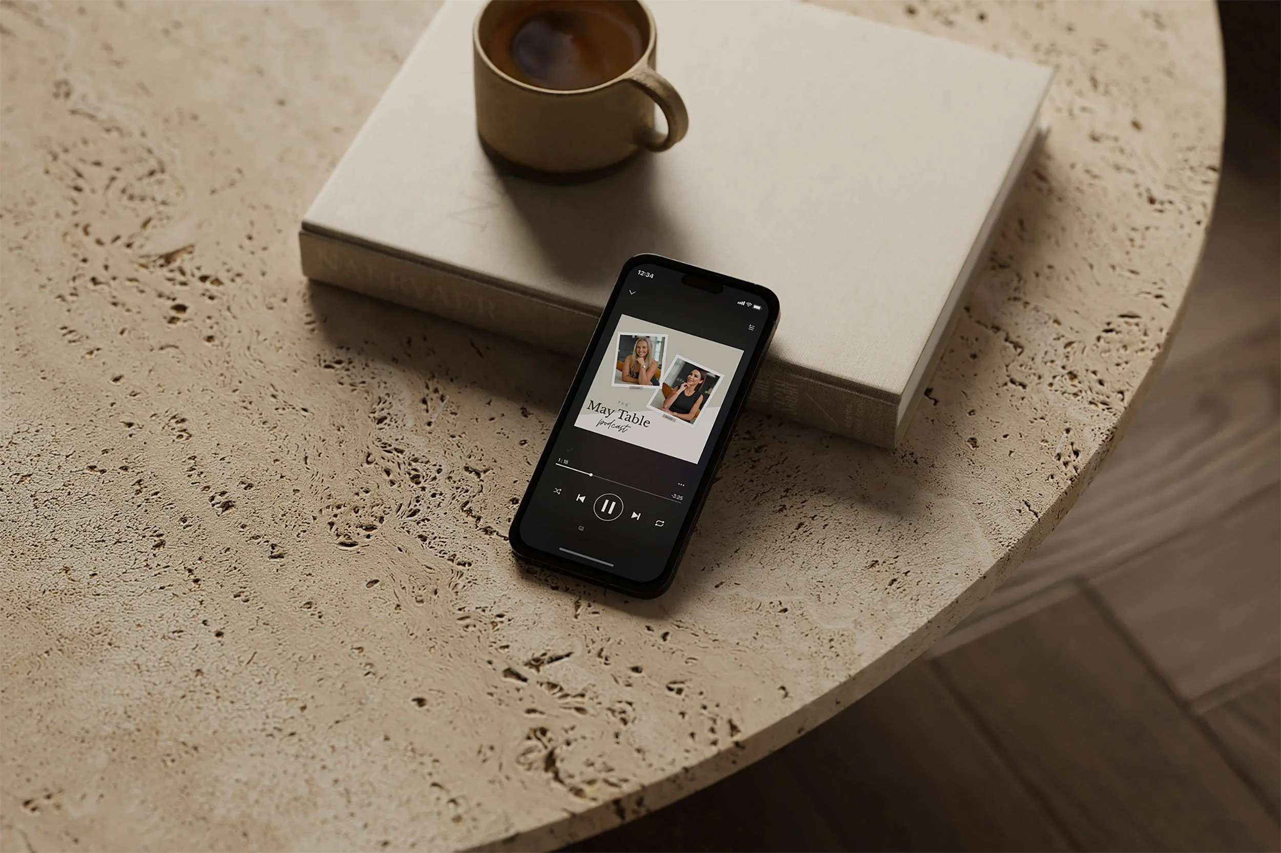 A smartphone placed on a textured beige table displays a playlist titled 'May Table podcast' with photos of two women. There is a closed book and a cup of coffee on top of it.