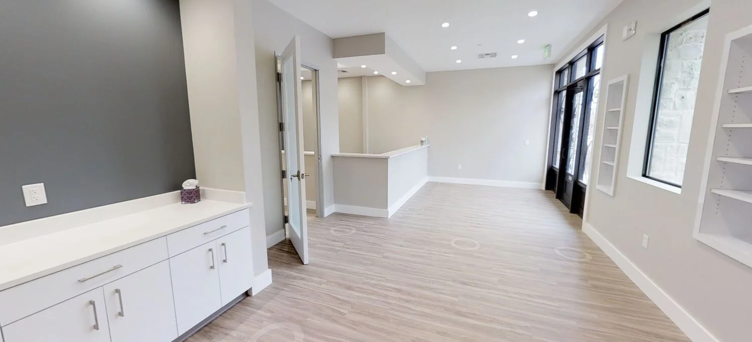 Empty commercial space with white and gray walls, large windows, light wood flooring, and built-in shelving.