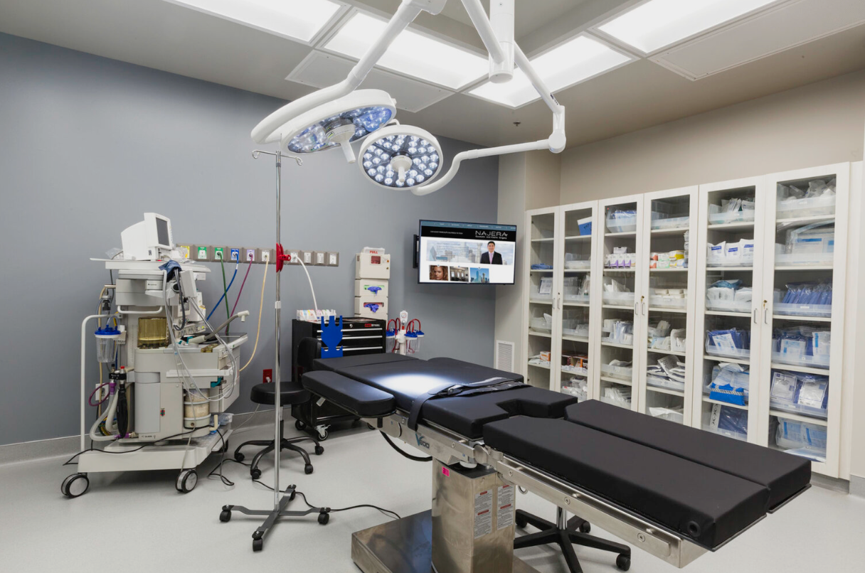An empty hospital operating room with a surgical table, medical equipment, overhead surgical lights, a monitor on the wall, and a storage cabinet filled with medical supplies.
