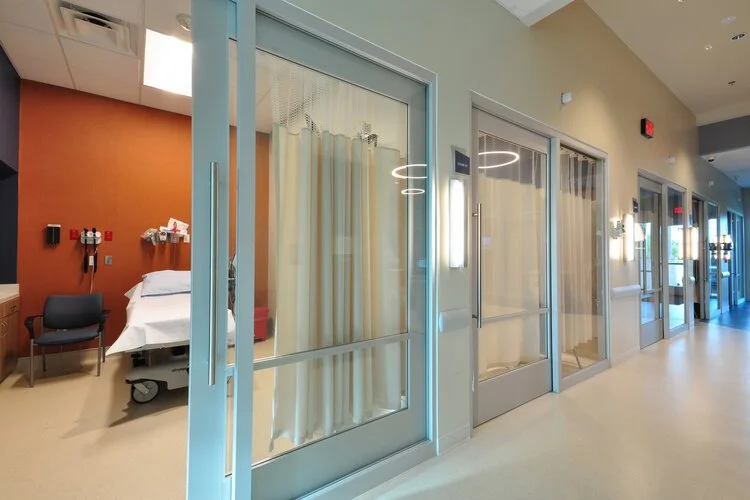 Hospital room with bed, medical equipment, and privacy curtains, visible through a glass door in a hospital corridor.
