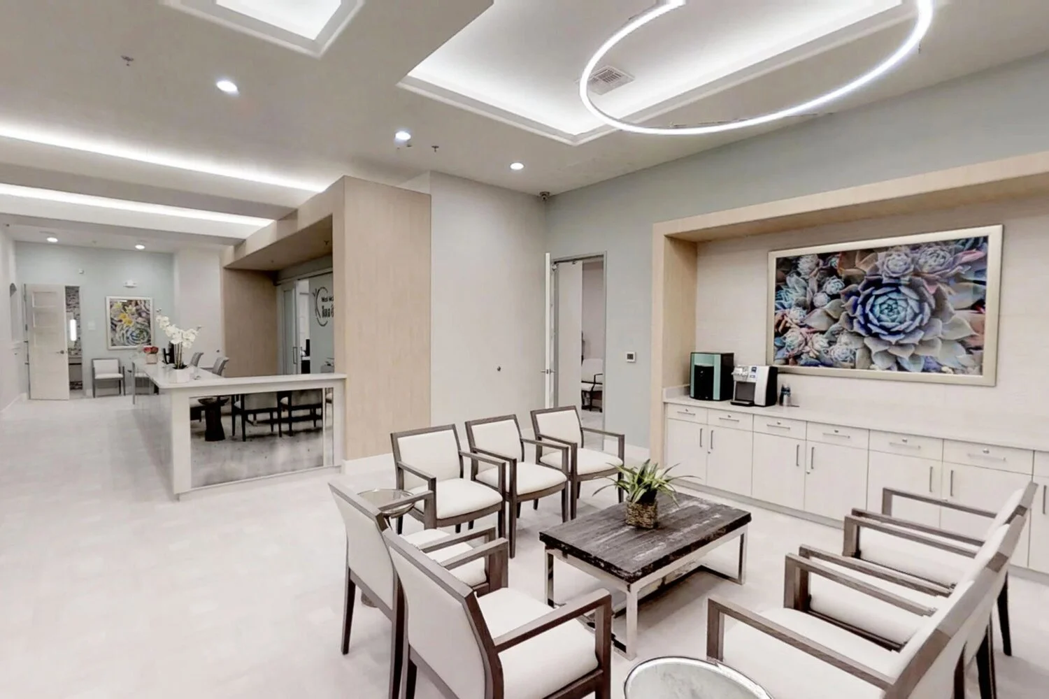 Modern waiting or lounge area with six white upholstered chairs around a dark wooden coffee table with a potted plant, light-colored walls, a large floral artwork above white cabinets, and a coffee station, with additional seating and a dining area visible in the background.