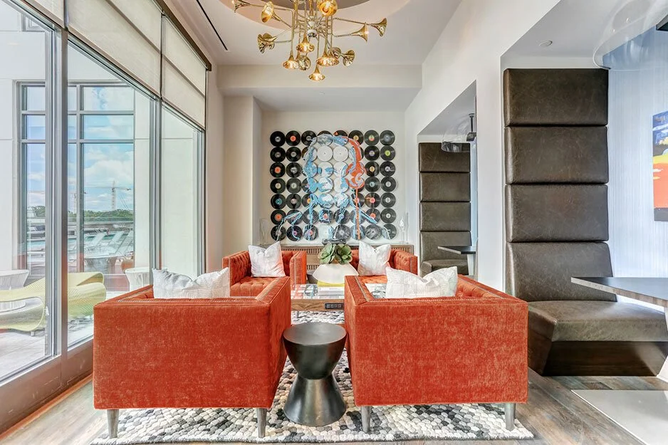 A modern living room with a large window, red sofas arranged around a coffee table, decorative pillows, a wall with a portrait made from vinyl records, and a chandelier hanging from the ceiling.