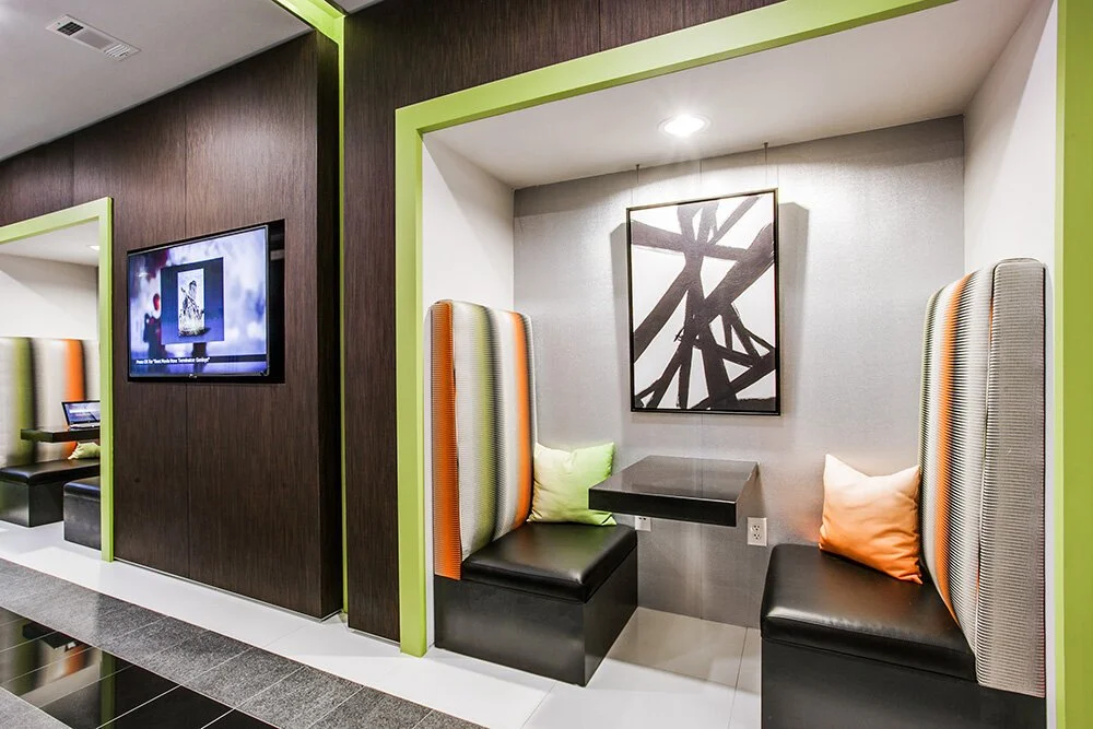 Two built-in booth seats with colorful high backrests and pillows, a small black table, modern artwork, and a wall-mounted TV in a contemporary hotel lobby.