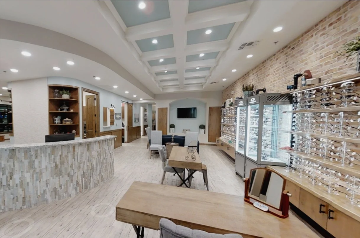 Interior of an optometry clinic or eyewear store with a display wall of eyeglasses, wooden furniture, and a reception area.