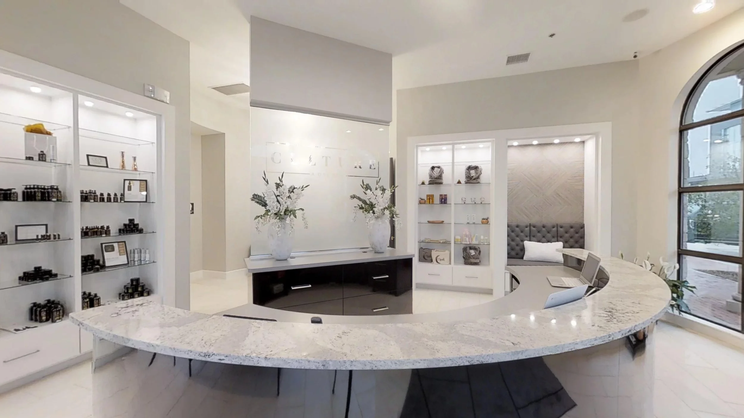 Hotel reception area with white curved marble desk, display shelves with products, floral arrangements, and seating nook near large windows.