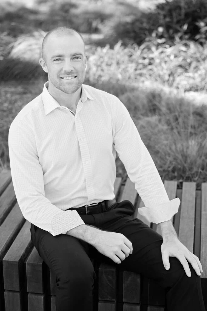 A man with a shaved head and beard crossing his arms, wearing a white dress shirt and dark trousers, sitting on a wooden bench outdoors, smiling at the camera.