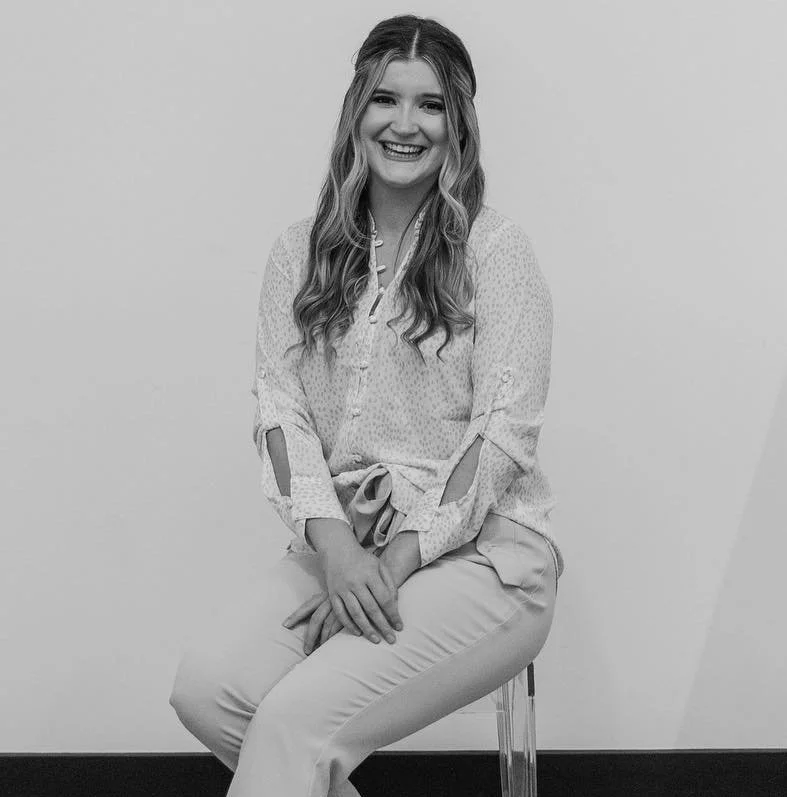 A woman with long wavy hair sitting on a transparent chair, smiling at the camera, wearing a blouse with cut-out sleeves and light-colored pants.