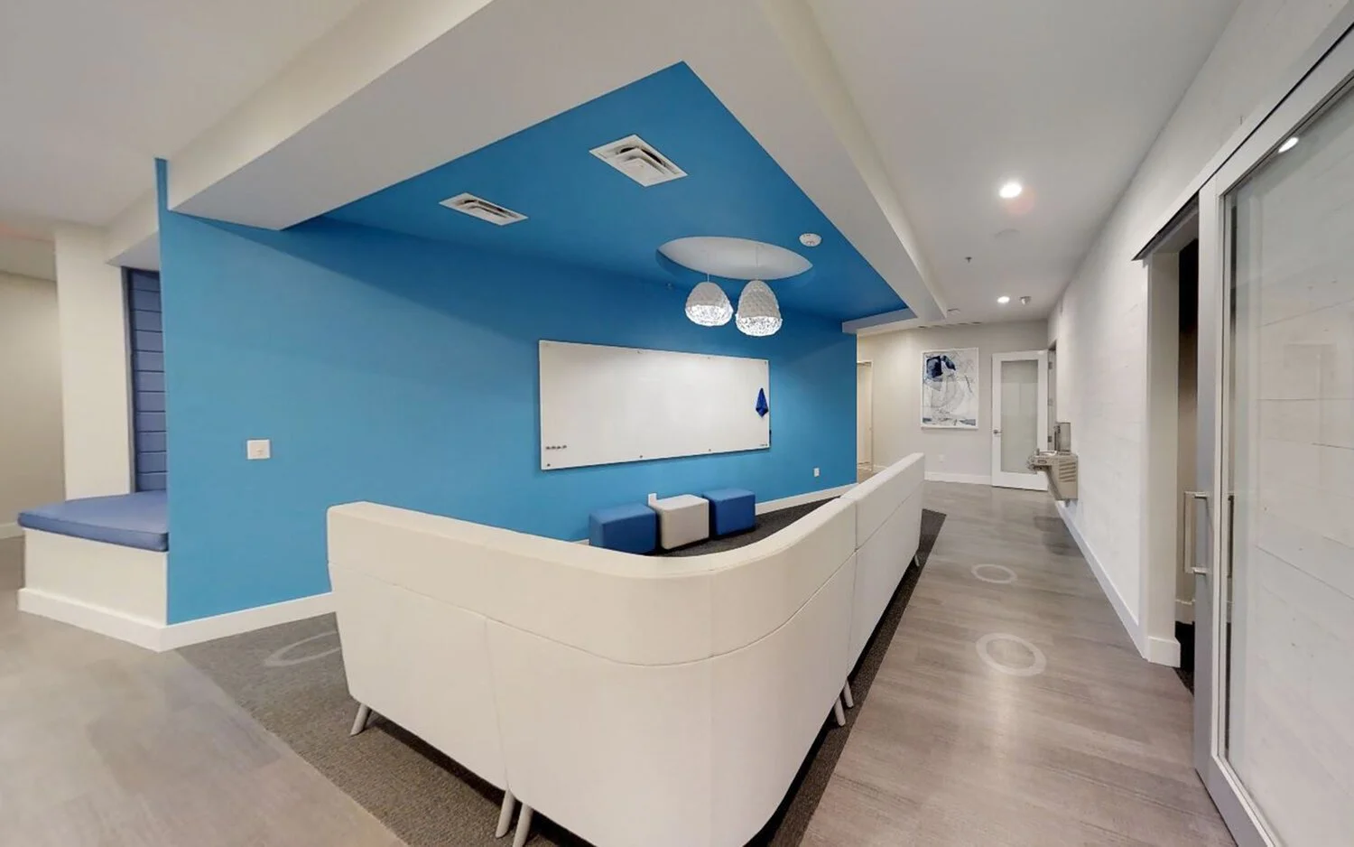 Modern waiting area with white curved sofa, blue accent wall, and whiteboard, in a well-lit corridor with wood flooring.
