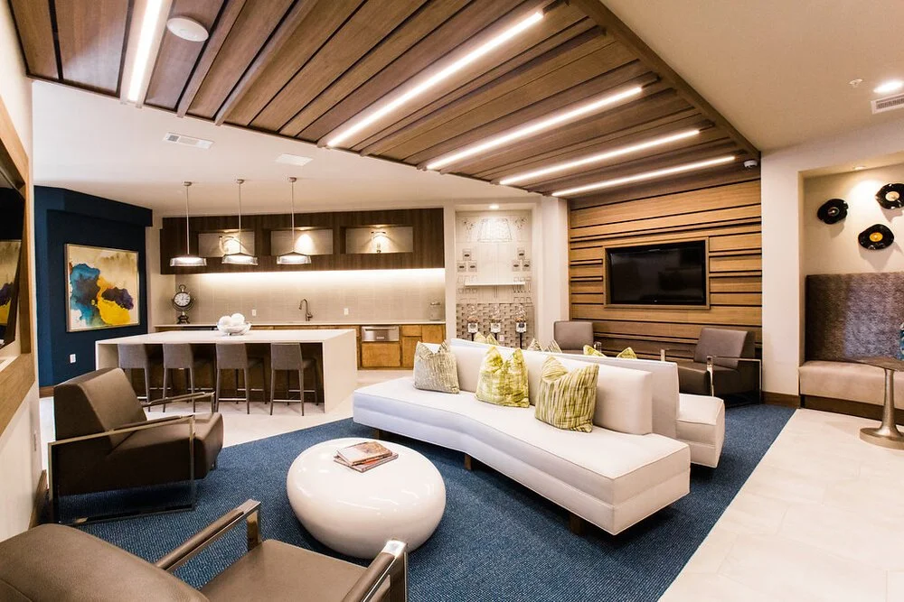 Modern living room with a kitchen in the background, featuring a white curved sofa with green pillows, a brown armchair, a round white coffee table, and a wooden ceiling with built-in lighting.