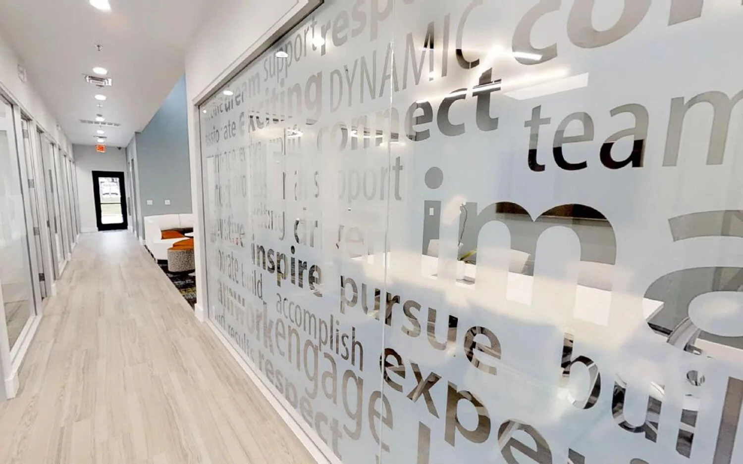 Modern office hallway with glass wall featuring motivational words and phrases, light wood flooring, and a seating area with orange cushions at the end.