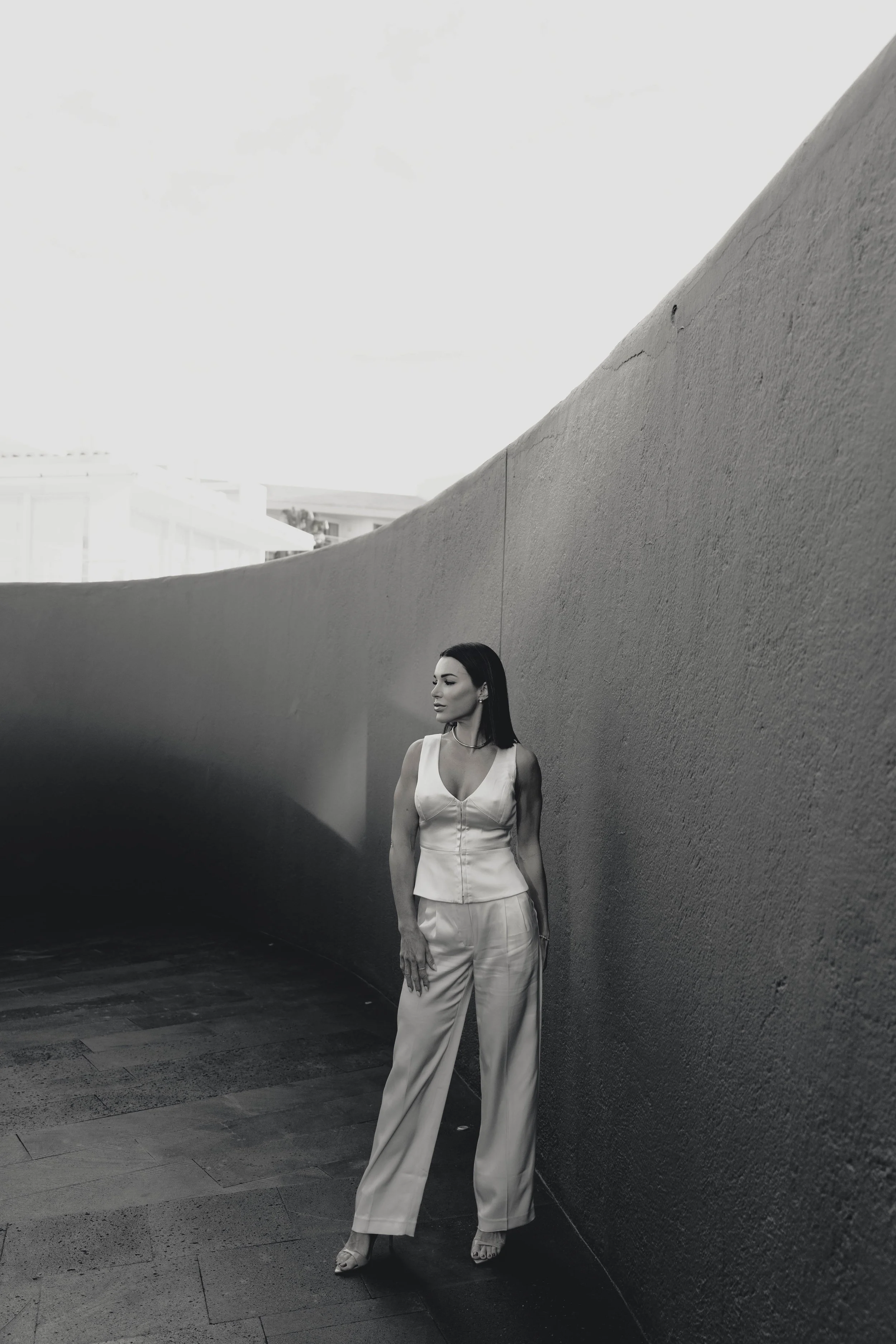 A woman in a sleeveless top and wide-leg pants stands against a curved concrete wall outdoors, with a bright sky above.