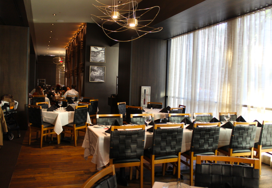 Interior of a modern restaurant with dark walls, wooden flooring, and large windows with sheer curtains; tables are set with white tablecloths and black napkins, surrounded by black leather chairs; there is stylish lighting and black and white wall art.