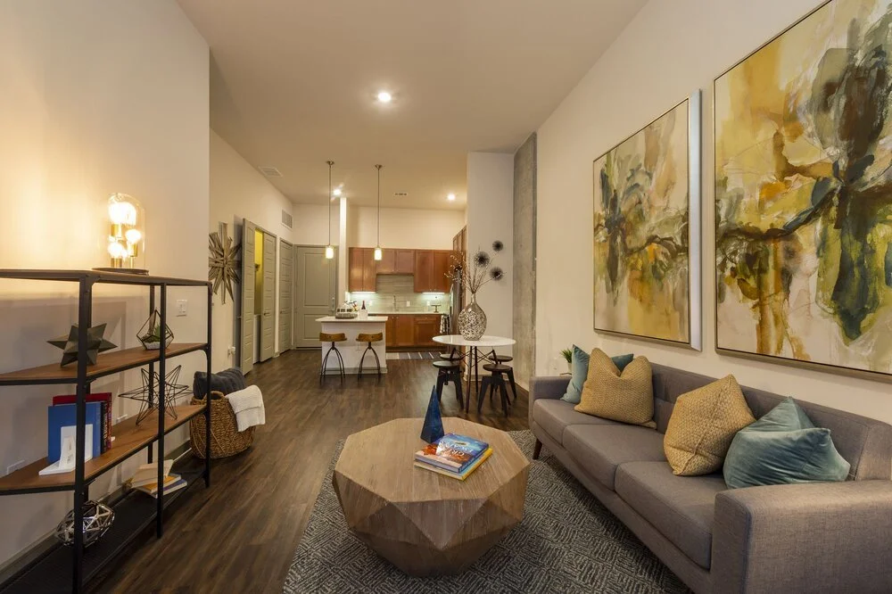 Modern living room with a gray sofa, colorful pillows, abstract wall art, a wooden coffee table with books, a black metal bookshelf, and an open kitchen with pendant lights.