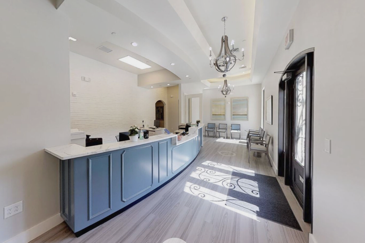 A reception area with a curved blue counter, chandelier, chairs along the wall, and sunlight casting shadows through an ornate glass door.