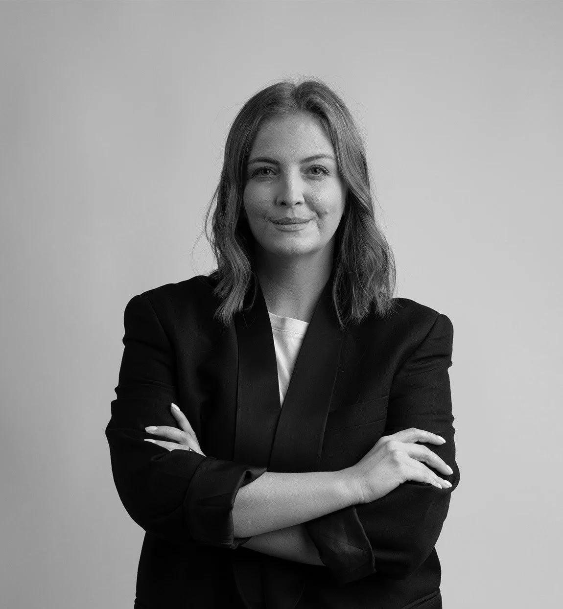 Black and white portrait of a woman with shoulder-length wavy hair, wearing a blazer over a light-colored top, standing with arms crossed in front of a plain background.
