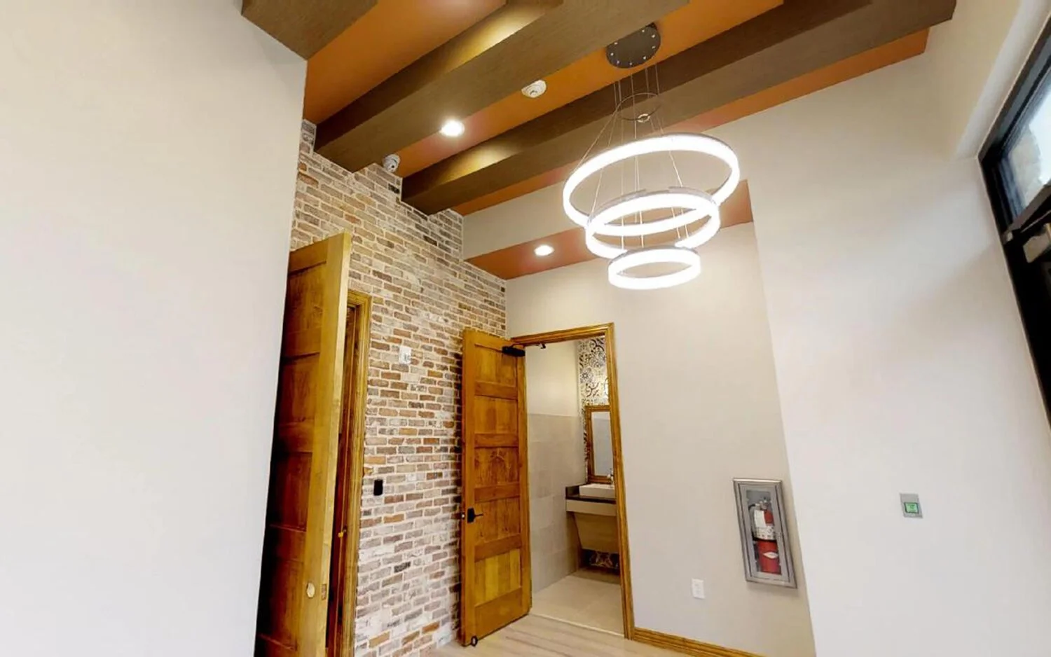 Interior view of a room with a brick accent wall, wooden doors, and a modern circular LED chandelier hanging from a ceiling with wooden beams. There is a small bathroom with a floating sink and mirror visible through an open door.