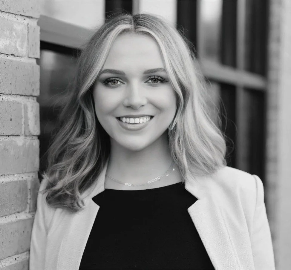 A smiling woman with wavy hair, wearing a blazer and a necklace, leaning against a brick wall outside building.