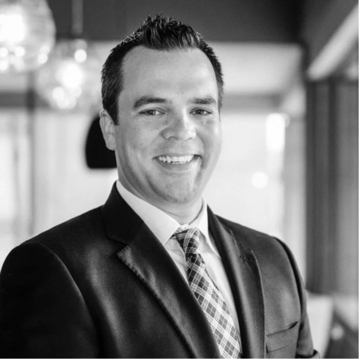 A smiling man in a suit, white shirt, and patterned tie, standing indoors with a blurred background.