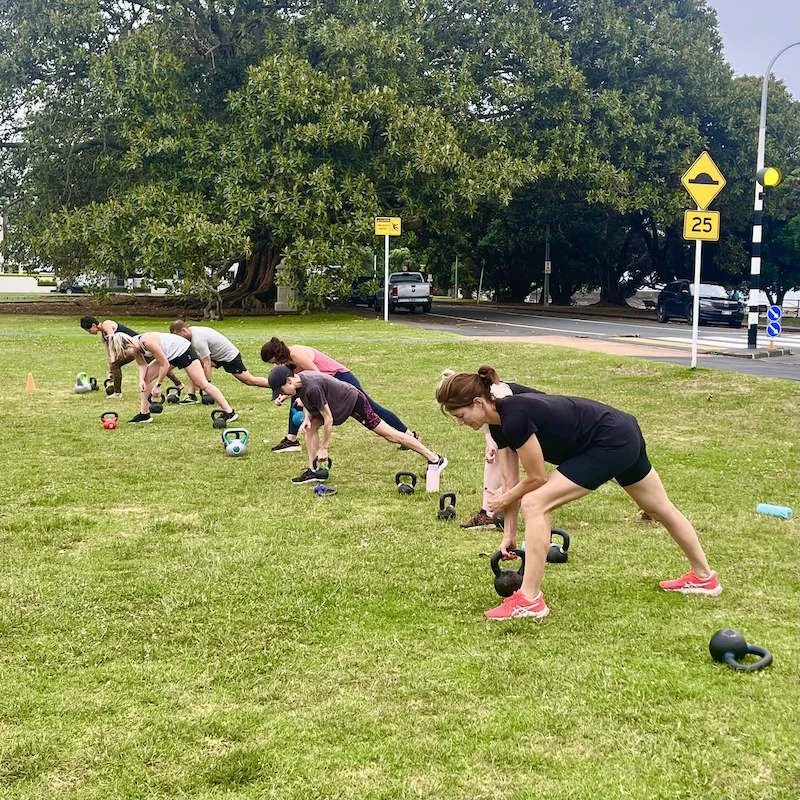 Six people, mostly women, exercising outdoors with kettlebells on a grassy field, with trees and a street in the background.