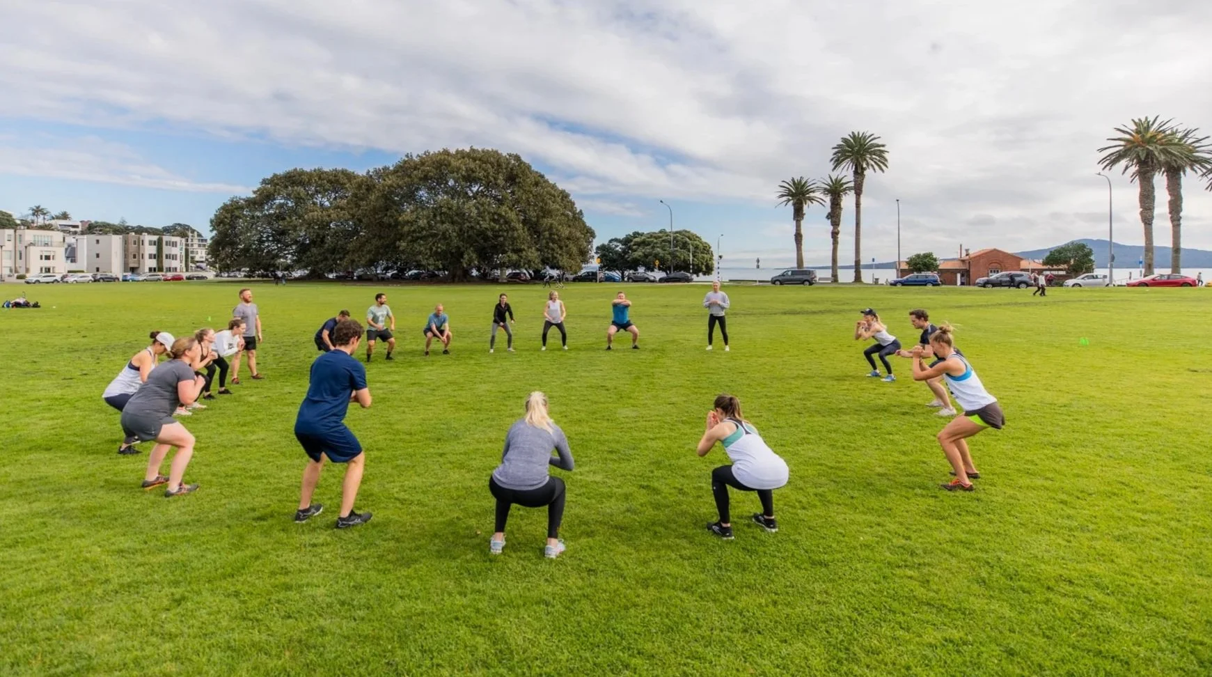 A group of people participating in a fitness class outdoors on a grassy field, with palm trees, cars, and buildings in the background under a partly cloudy sky.