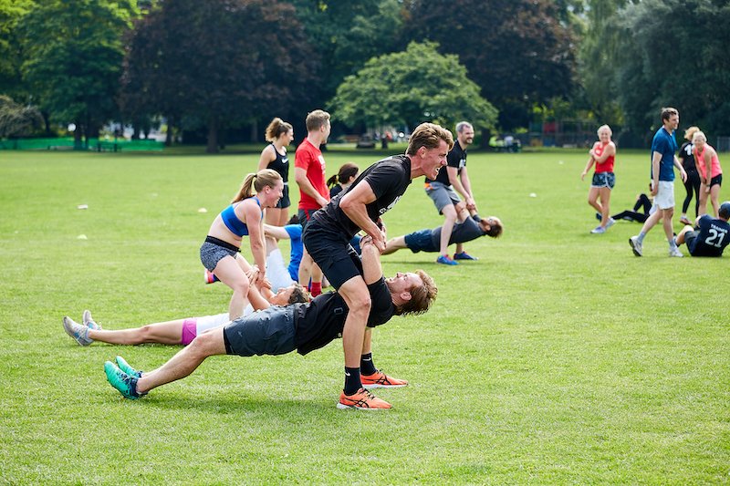 Two men working out in a park, with the man in black shirt balancing on the shoulders of the other. There are children and adults in the background, some stretching and some watching.