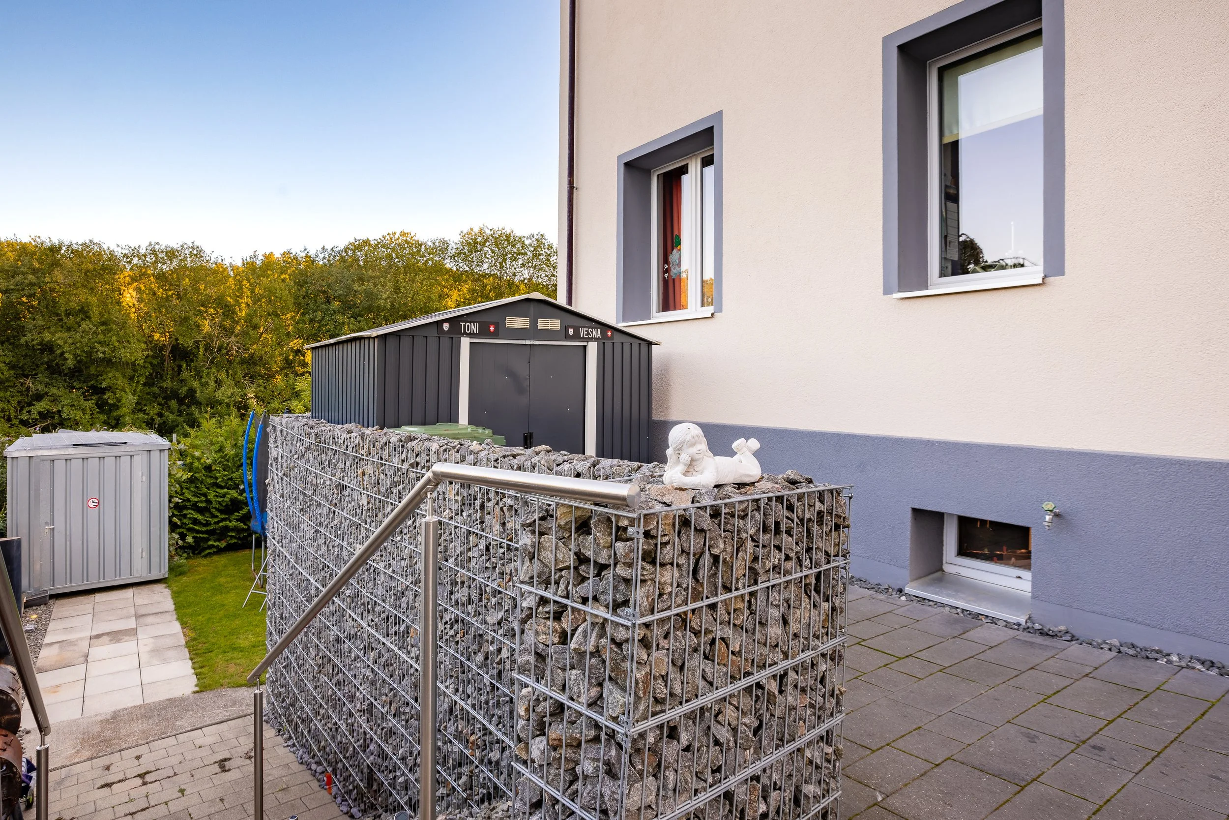 Garten mit einem hohen Gabionenzaun, einem kleinen Hilfsgebäude, einem Kinder-Gartenspielzeug, einer weißes Kinderskulptur, einem Haus mit sichtbaren Fenster und Garten. Im Hintergrund Bäume und blauer Himmel. Haus in Neuhausen kaufen bei FREITAG IMM
