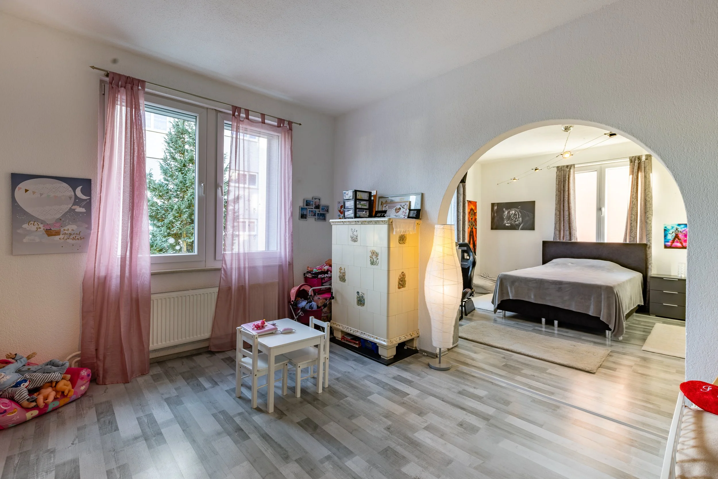 Ein offener Raum, der in einen Spielbereich für Kinder und ein Schlafzimmer unterteilt ist, mit großen Fenstern, pinken Vorhängen, einem kleinen weißen Tisch, einem Kinderbett, und einem großes Bett im Schlafzimmerbereich.
