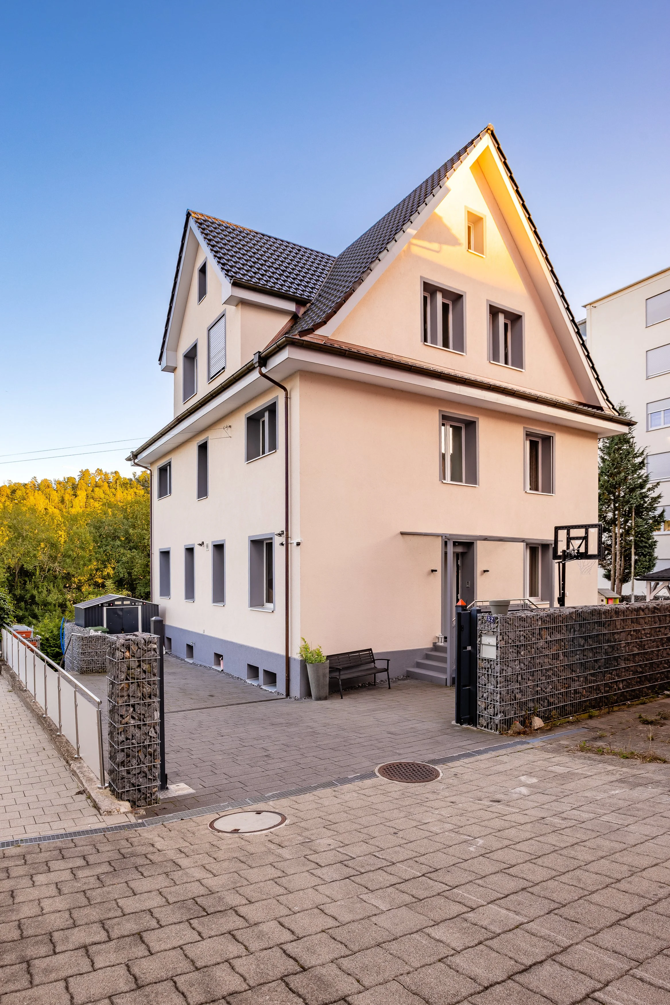 Ein mehrstöckiges, modernes Haus mit hellem Putz und dunklem Dach, umgeben von einem gepflasterten Hof und einem Zaun aus Gabionen. Im Hintergrund sind Bäume und ein Himmel mit Sonnenlicht.