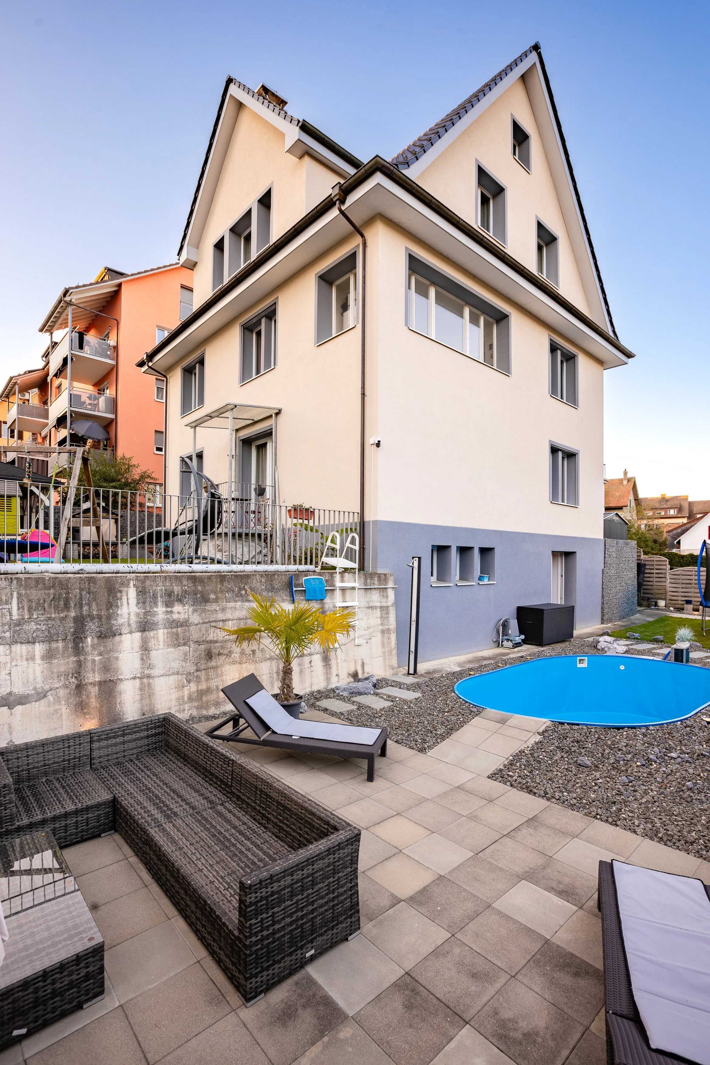 Geräumiger Garten mit Swimmingpool, Sonnenliege, Rattan-Sitzmöbeln und kleinen Palmen, im Hintergrund ein Mehrfamilienhaus mit mehreren Fenstern.