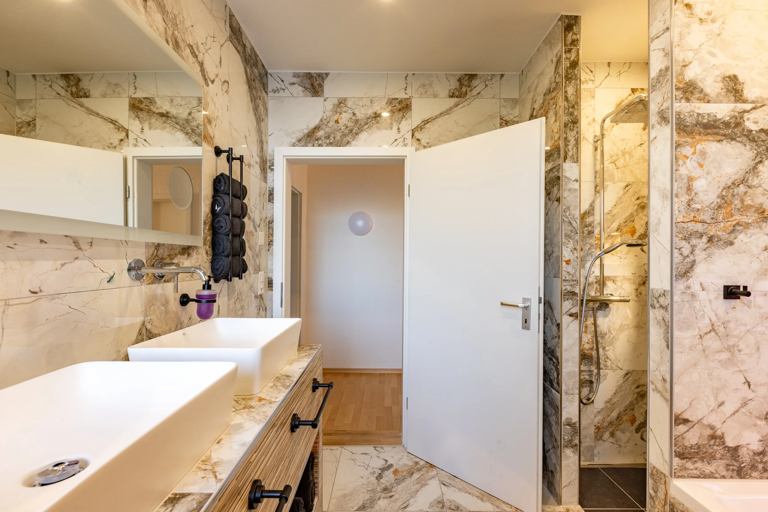 Ein Badezimmer mit Marmorkacheln, Doppelwaschbecken, einem Spiegel, einem Badetuchhalter mit schwarzen Handtüchern und einer begehbaren Dusche.