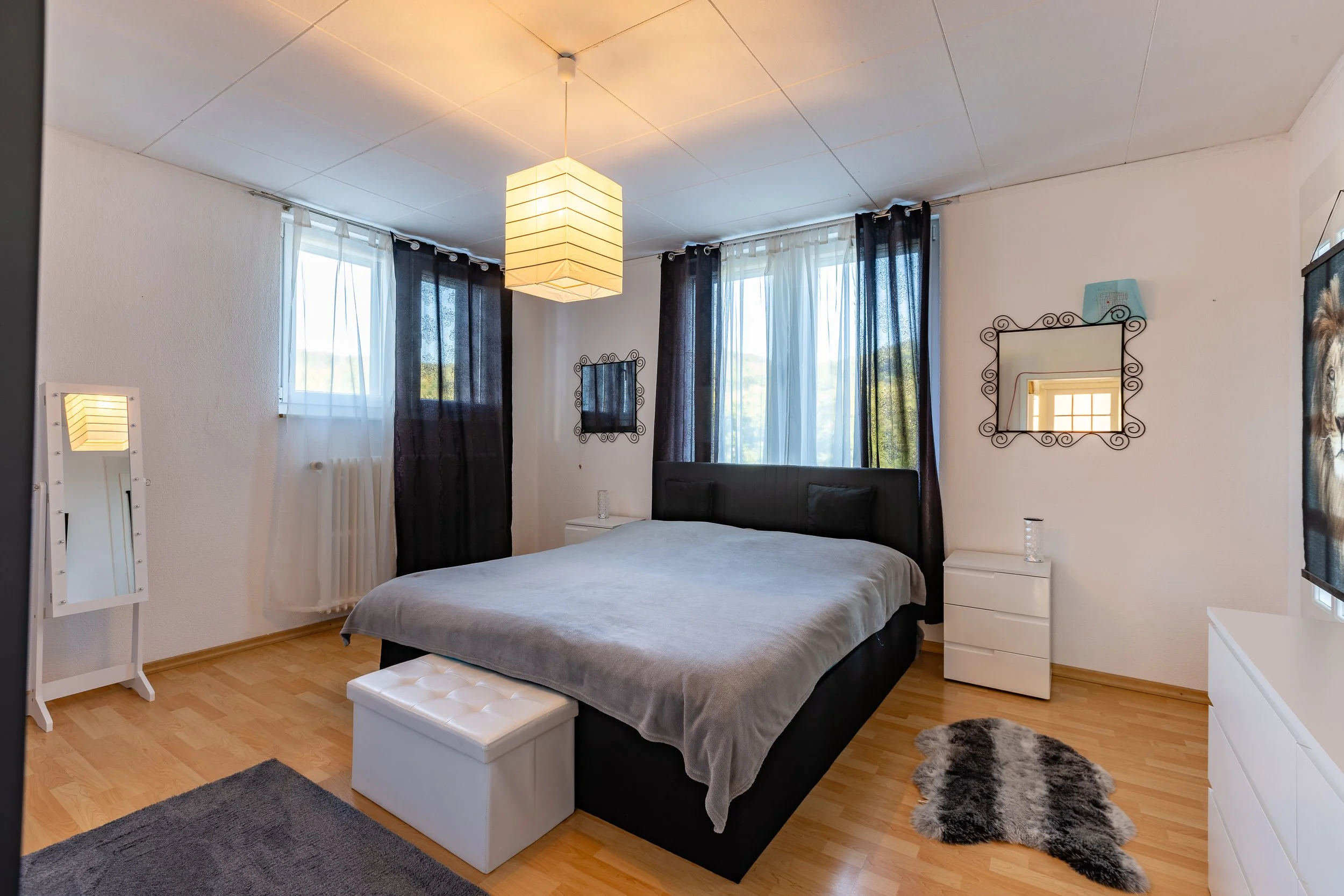 Schlafzimmer mit Doppelbett, schwarzen Kopfteil, weißen Nachttischen, schwarze Vorhängen, weißen Vorhängen, großen Fenstern, einem bemalten Spiegel, einem kleinen Stehspiegel, einem weißen Hocker am Bett, einem hellen Lampenschirm an der Decke, einem