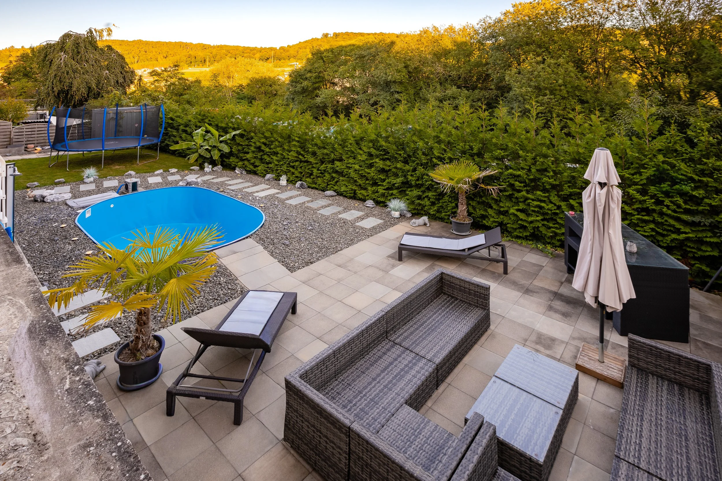 Ein moderner Garten mit einer kleinen blauen Pool, Sonnenliegen, einem Sonnenschirm, einem Trampolin und viel Grün, umgeben von Bäumen und einem Zaun. Haus in Neuhausen kaufen bei FREITAG IMMO: www.freitagimmo.ch