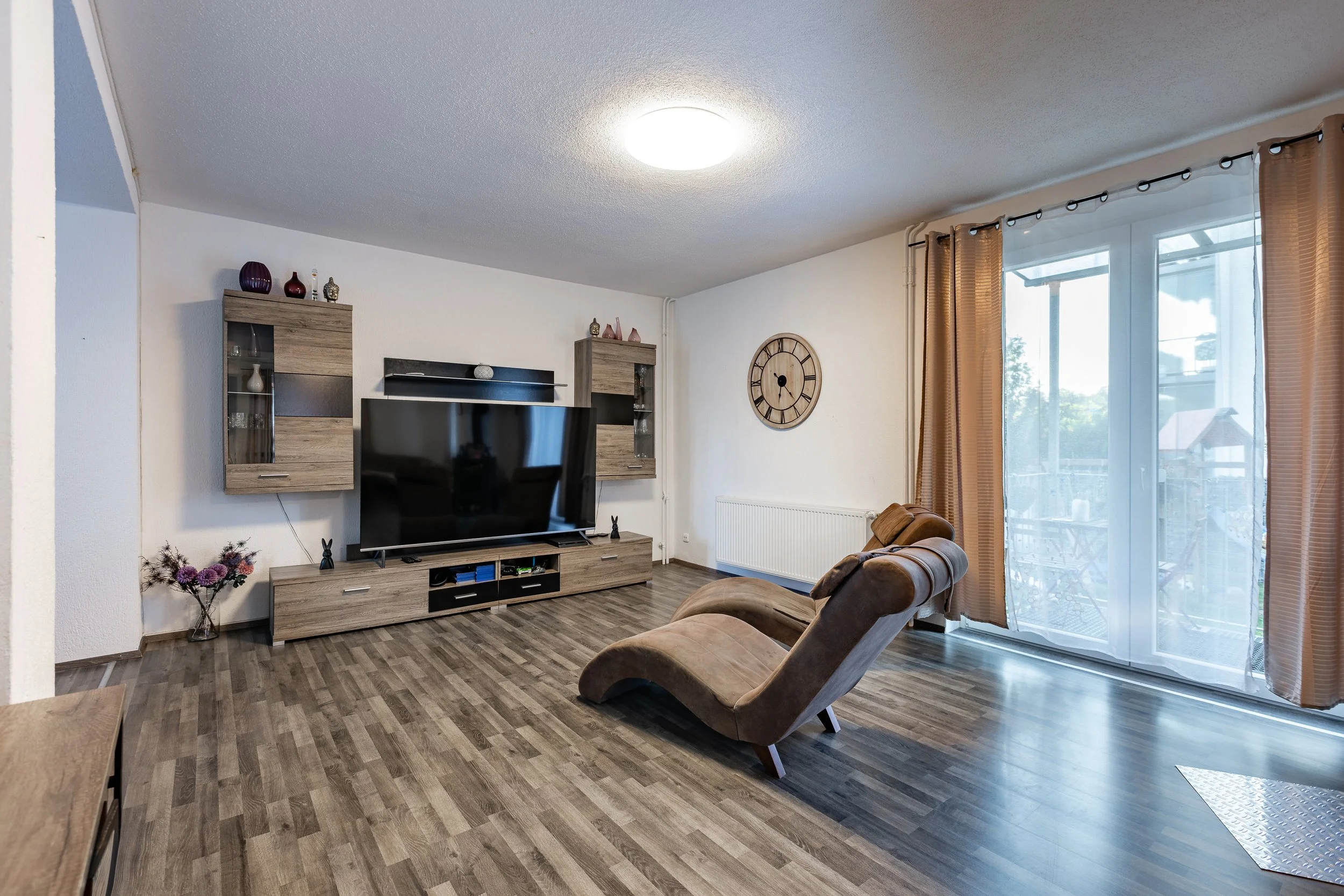 Wohnzimmer mit Fernseher, Wandregalen, Couchtisch, Sesseln, Vorhängen und Fenster mit Balkon