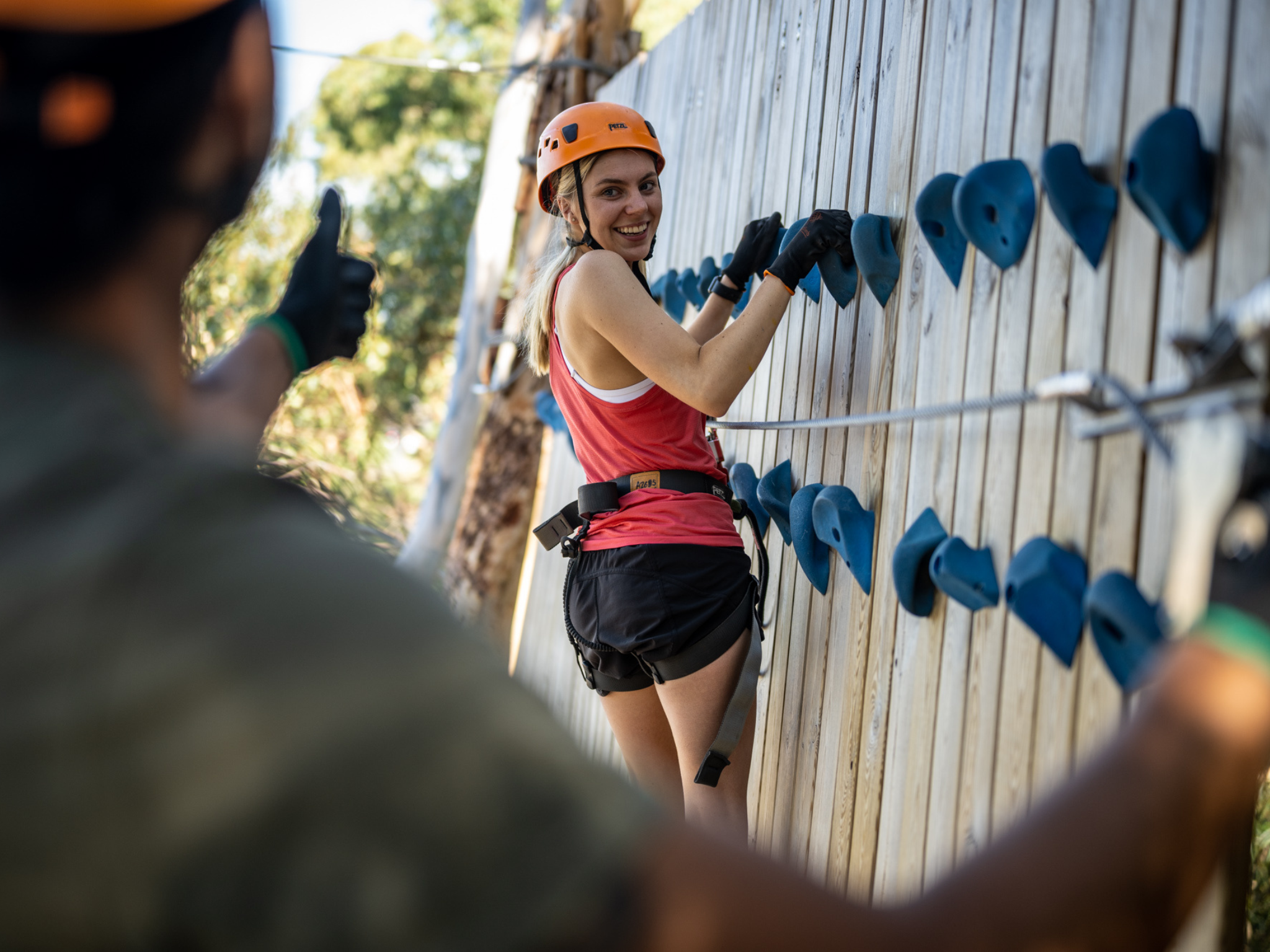 Woman rock climbing on outdoor wall with blue holds, wearing safety harness and helmet, giving a thumbs up; another person blurred in foreground.