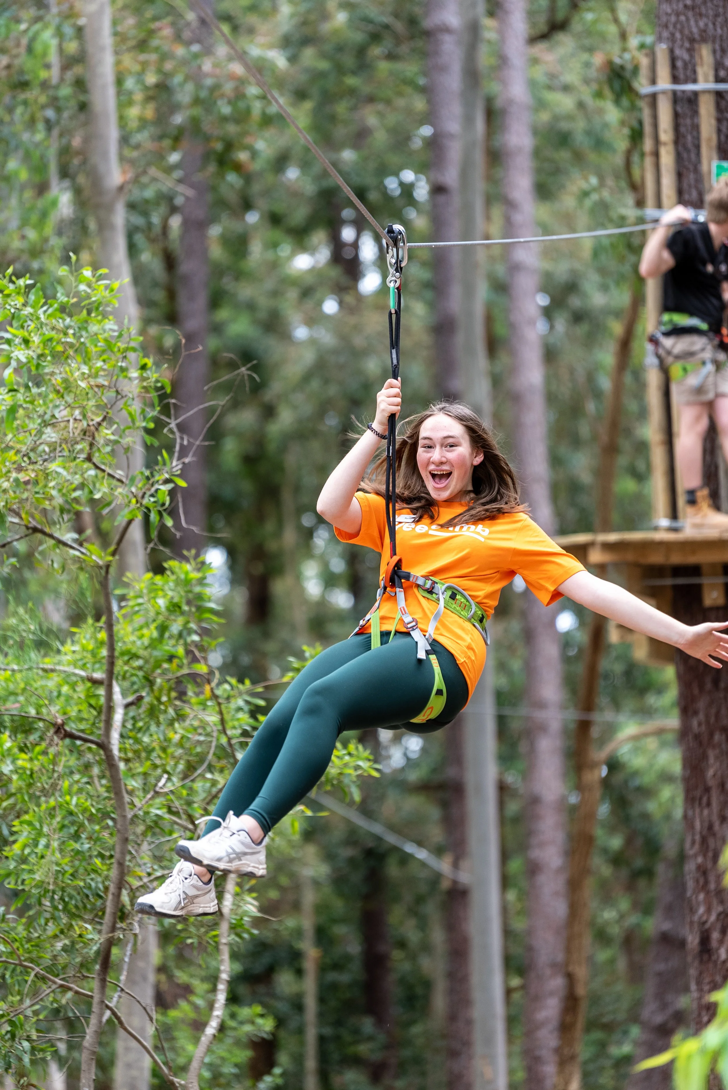 A person wearing an orange shirt and green leggings ziplining through a forested area with a joyful expression on their face.