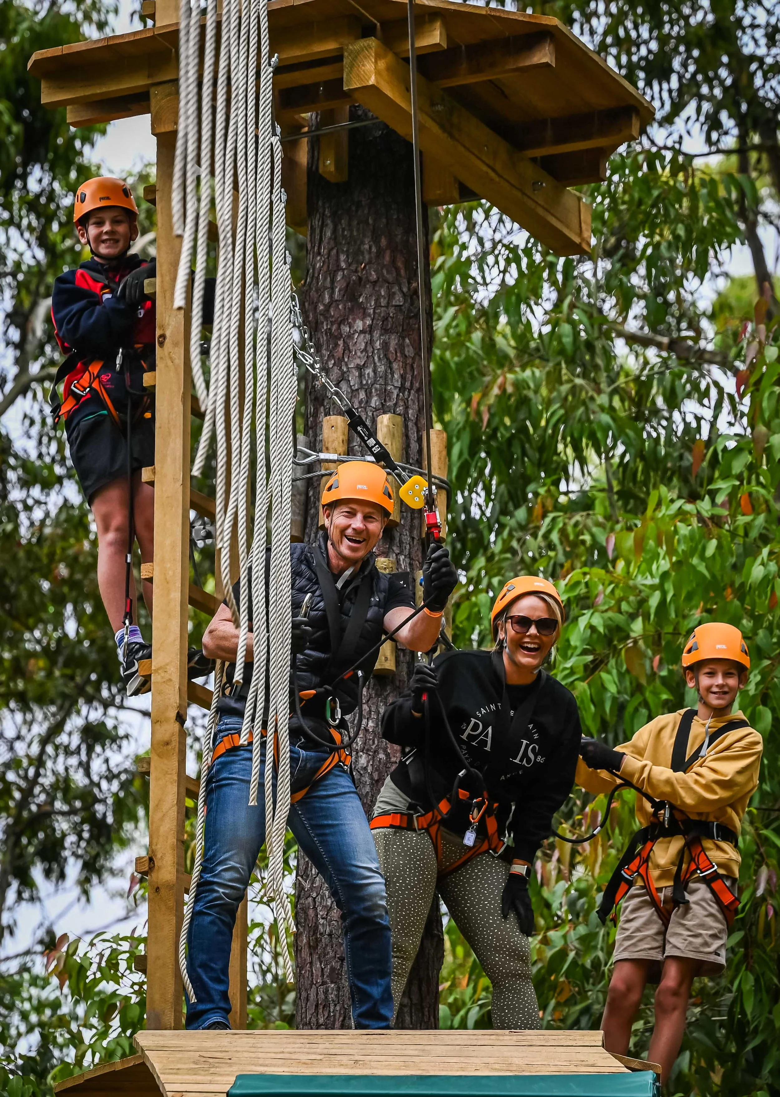 Group of people on a zip line platform in a forest, wearing helmets and safety harnesses.