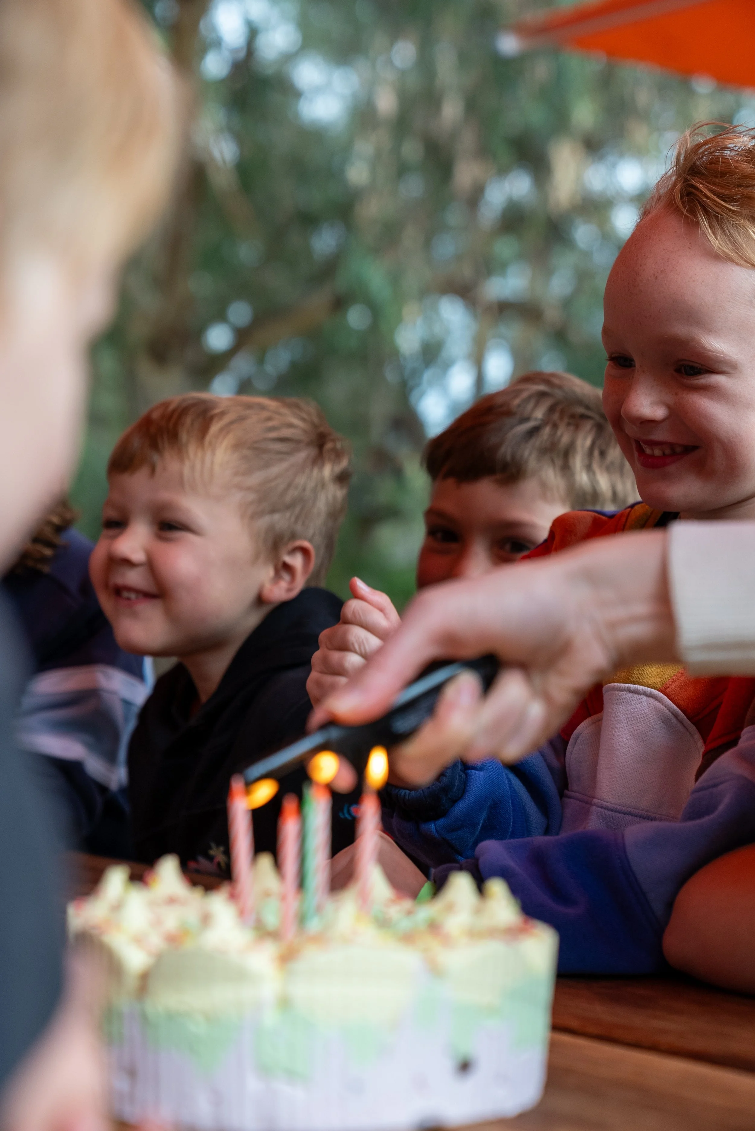 Group of children smiling as candles on a birthday cake are lit outdoors.