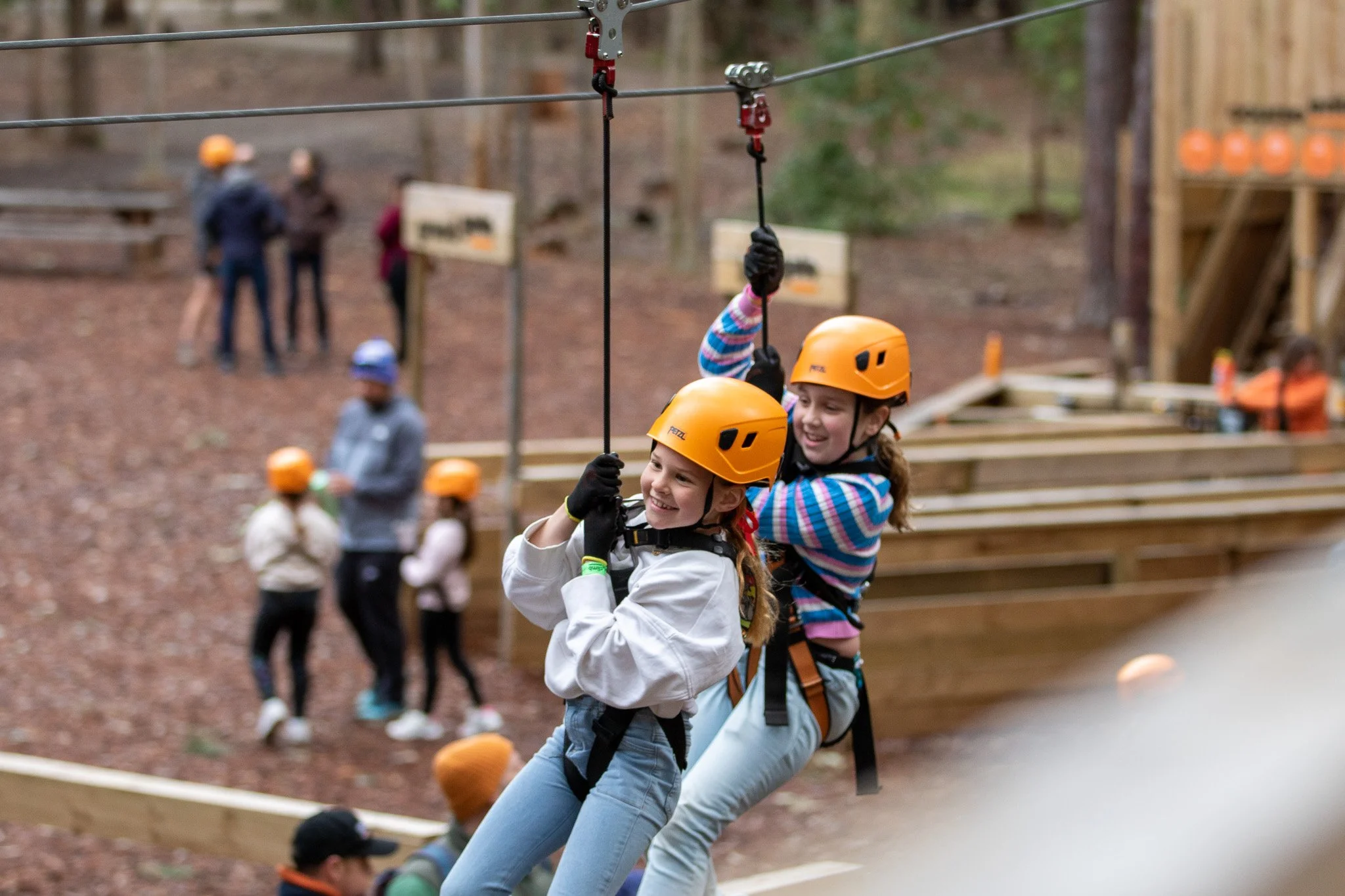 Two children wearing helmets and harnesses enjoy ziplining at an outdoor adventure park.