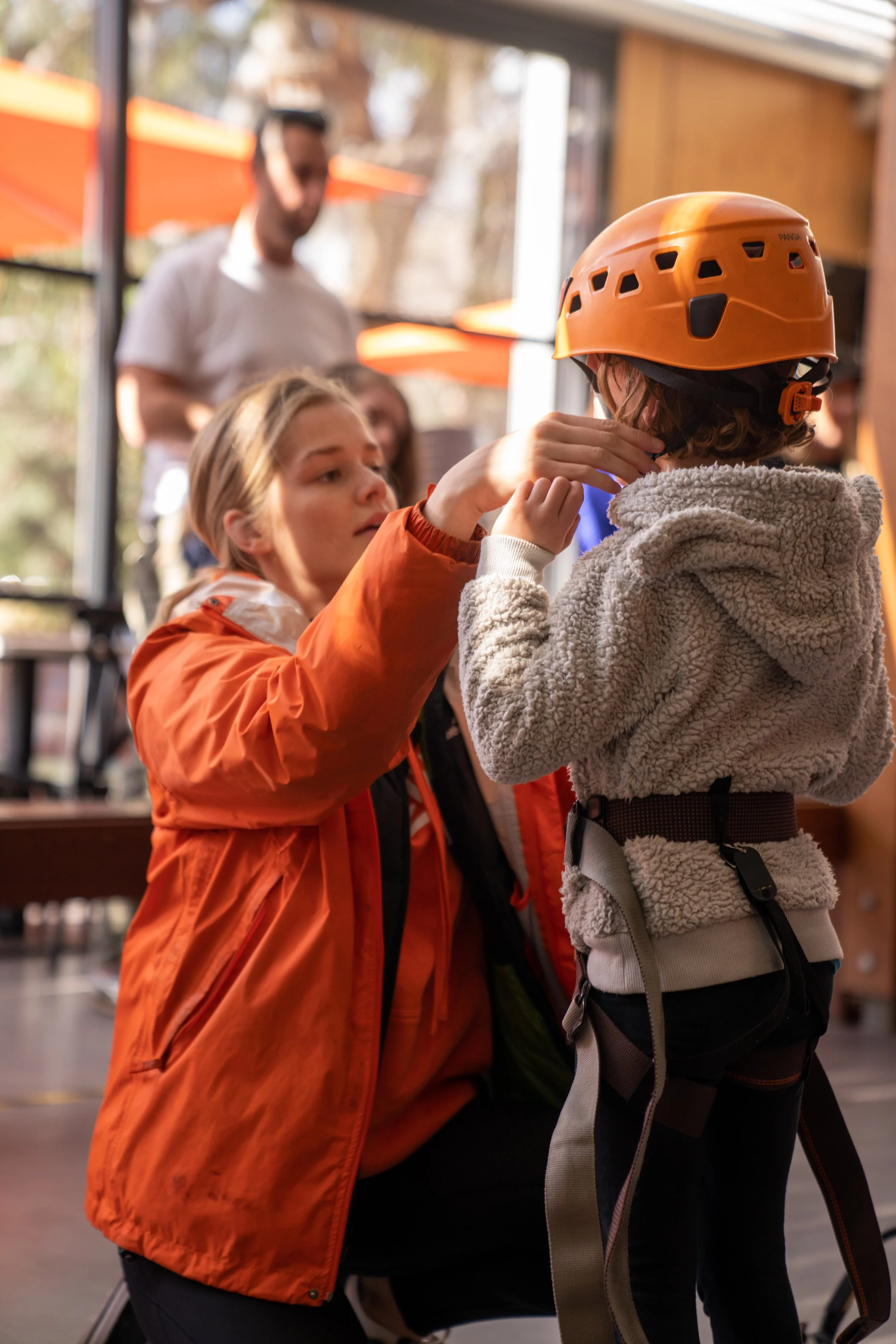 A woman in an orange jacket assists a child wearing a helmet and climbing harness indoors.
