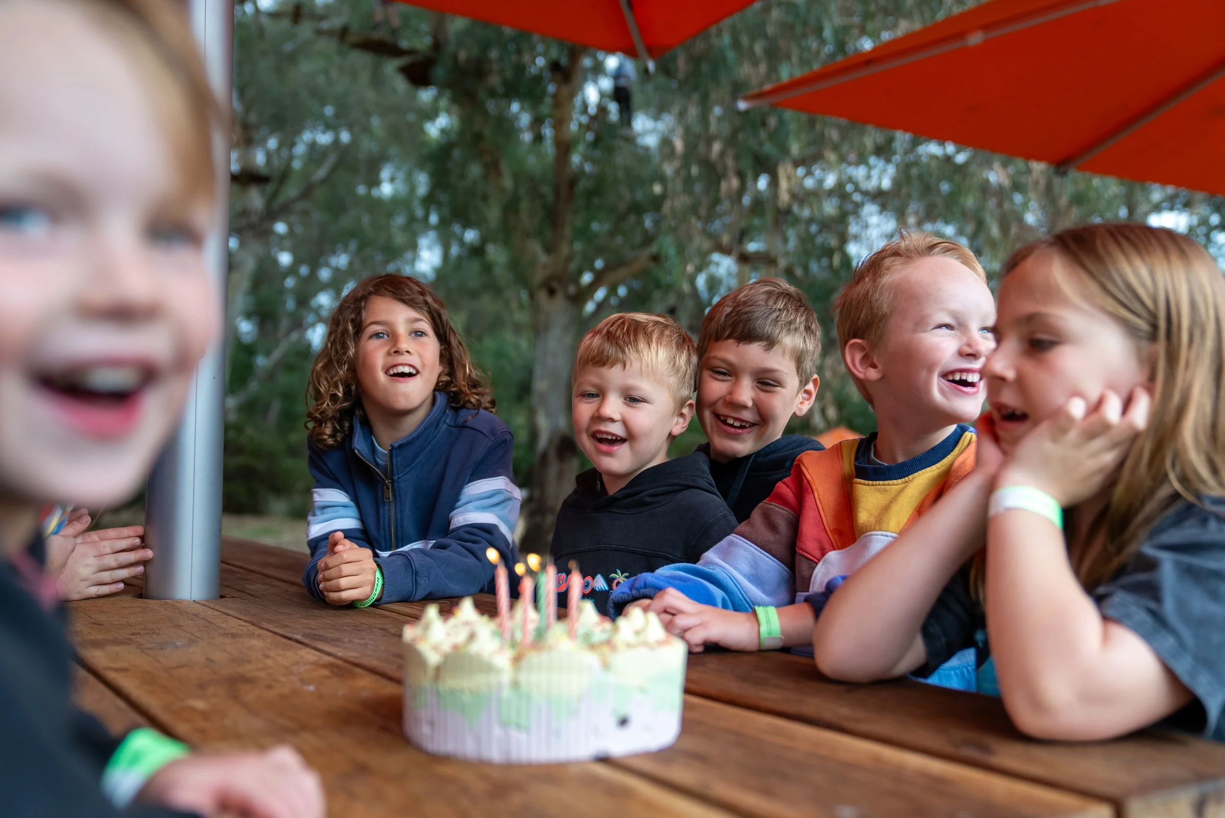 Group of children smiling at a birthday party, sitting outside with a cake on the table.