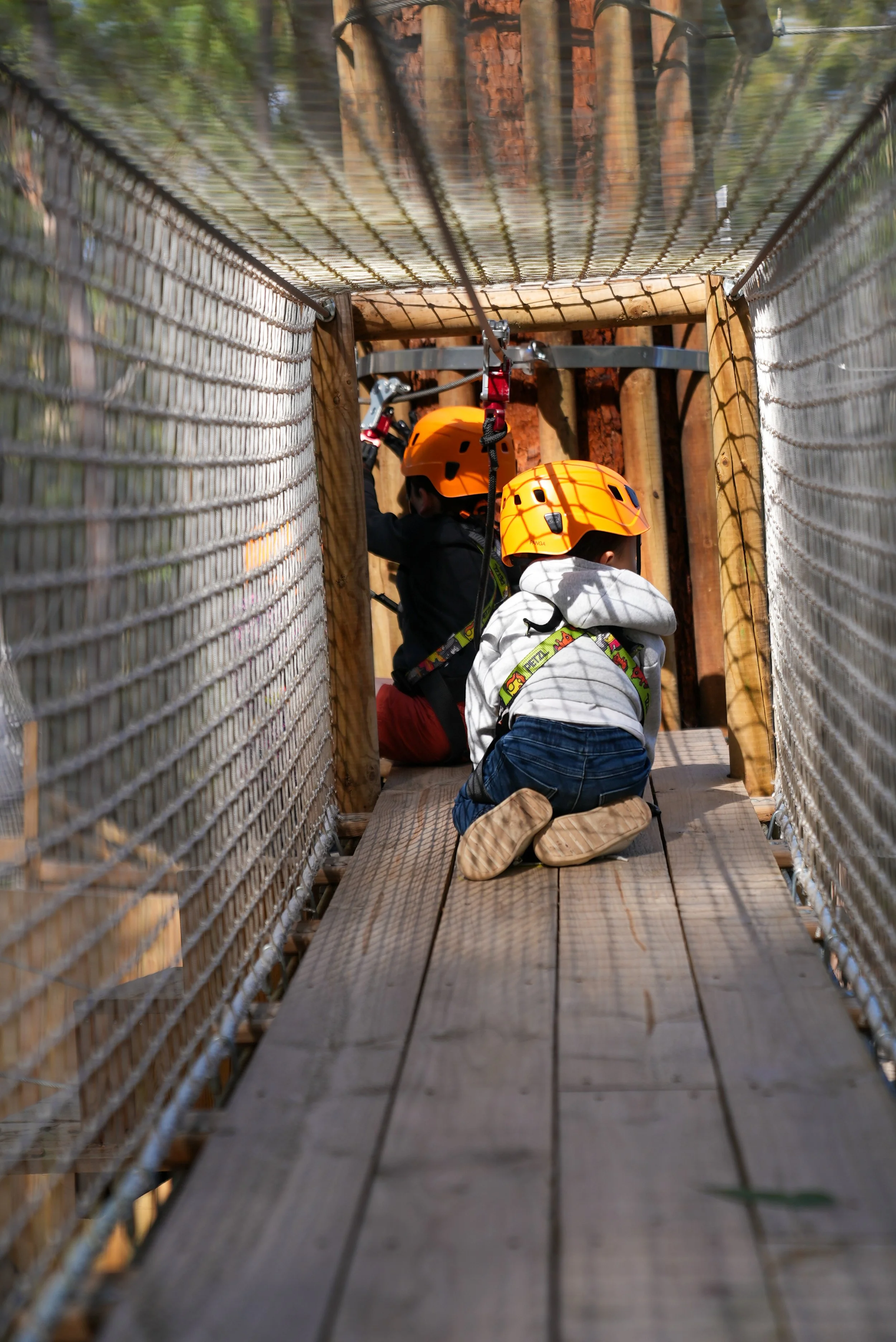 Two children in orange helmets crawling through a wooden adventure park bridge, secured with harnesses and safety ropes.