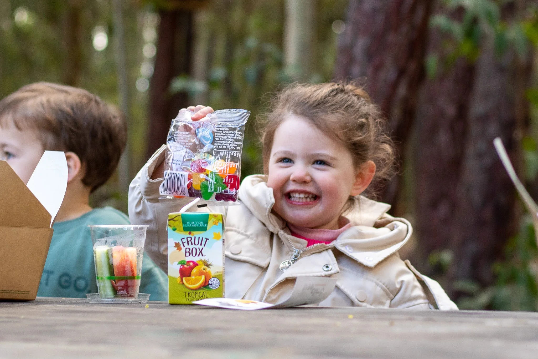 A smiling child holding a bag of colorful gummy candies, sitting at a table with a carton of fruit juice and a cup of sliced fruit outdoors.
