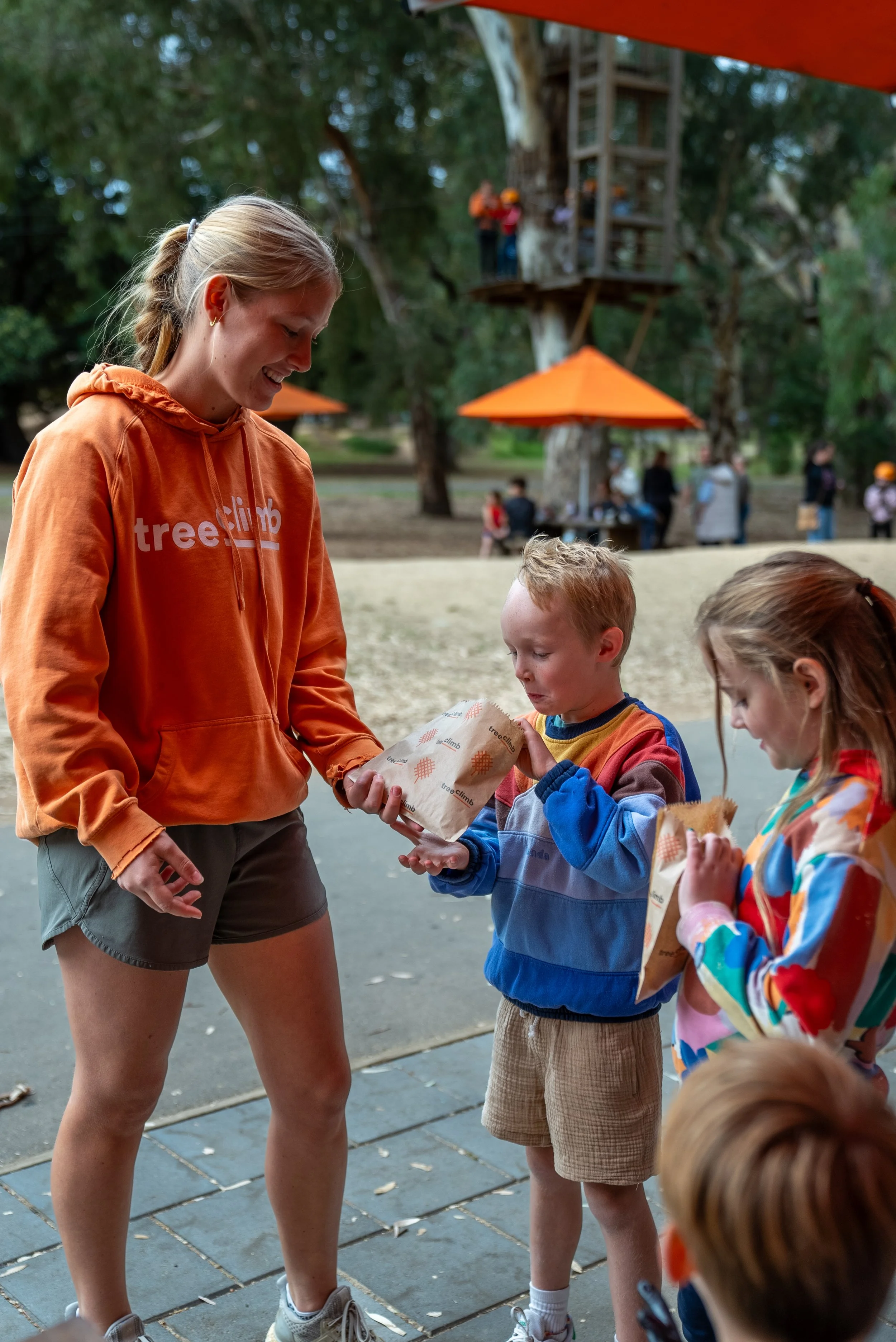 Woman in orange hoodie gives snacks to children in a park setting with orange umbrellas and trees in the background.