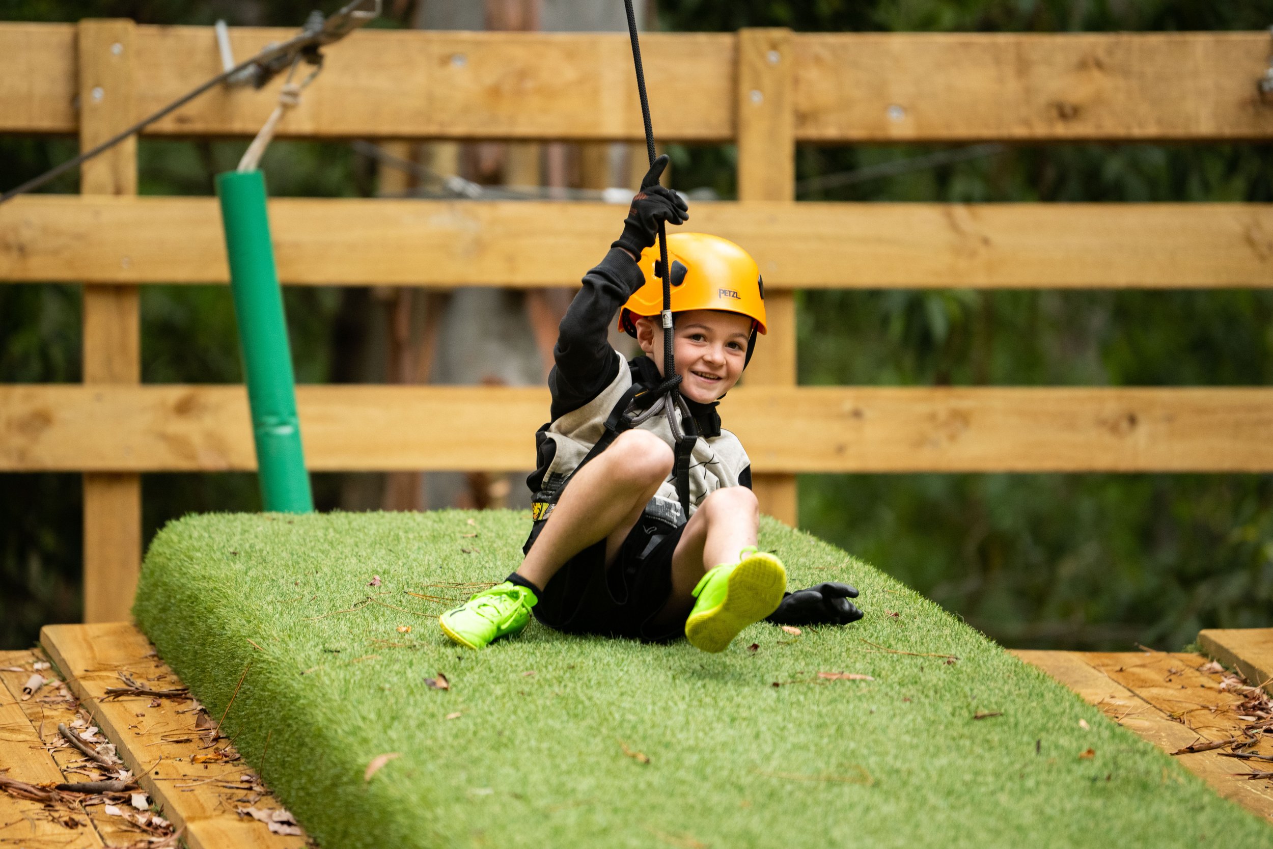 Young boy on a zip line, wearing a helmet and gloves, sitting on a grass-covered platform.